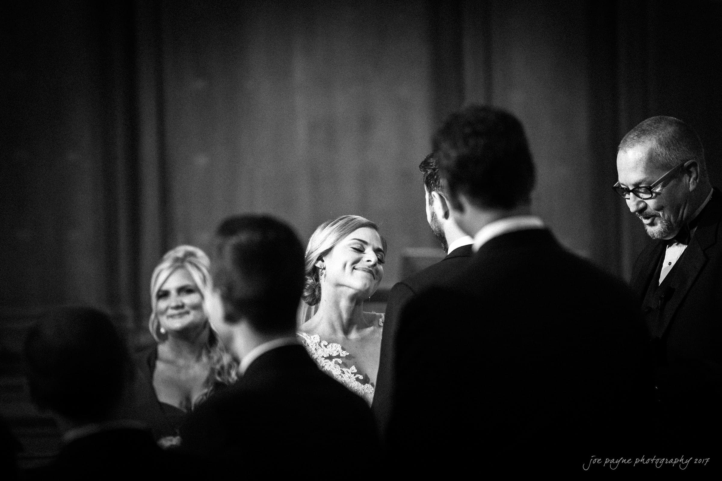 A black and white photograph of a wedding ceremony. A bride smiles at her groom, surrounded by a small group of people. An officiant stands nearby. The scene is intimate and joyful.