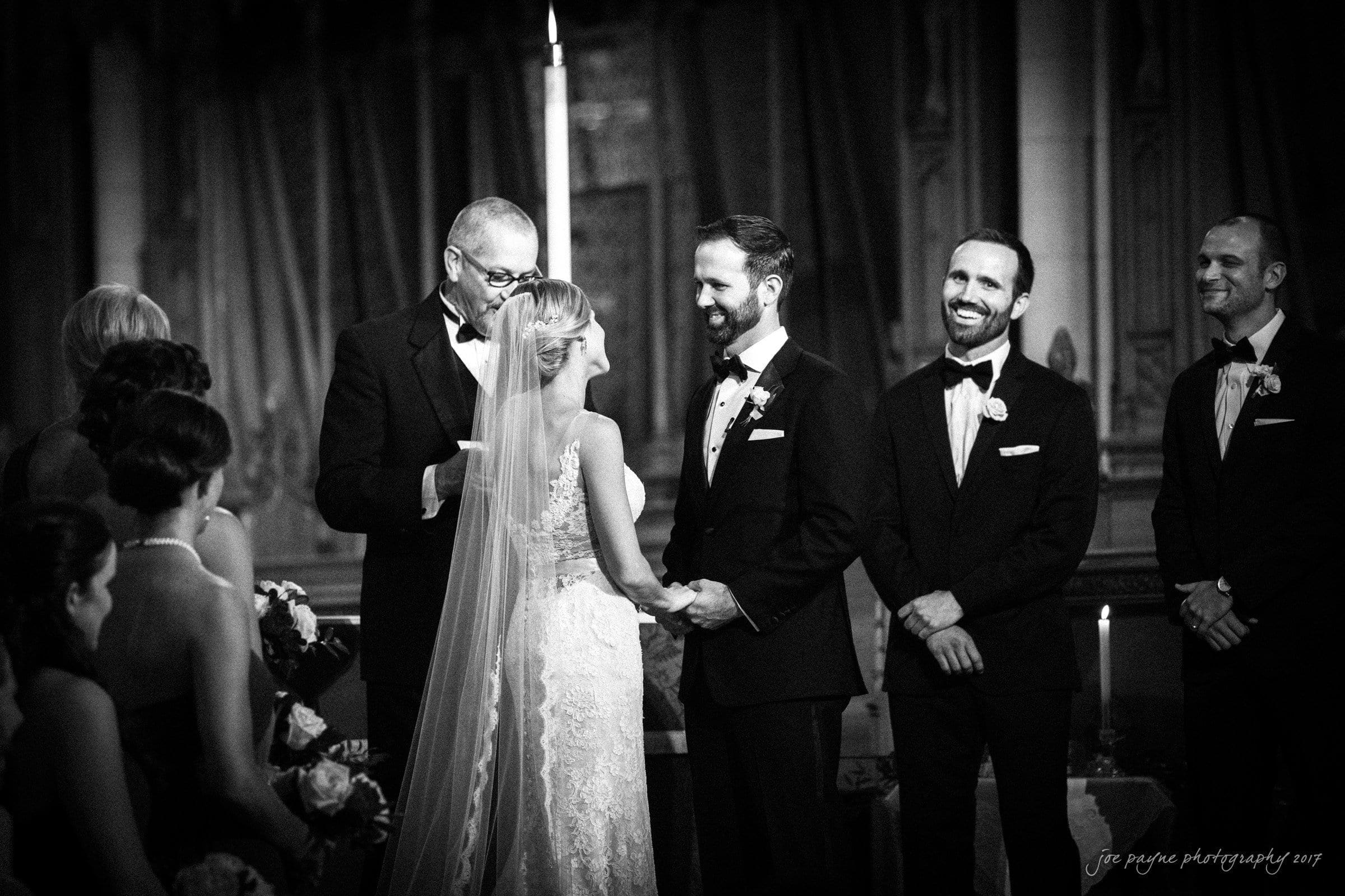 A black-and-white photo of a wedding ceremony. The couple is smiling at each other, holding hands. The officiant and two groomsmen stand beside the groom. Bridesmaids are visible, holding bouquets. A candle glows in the background.
