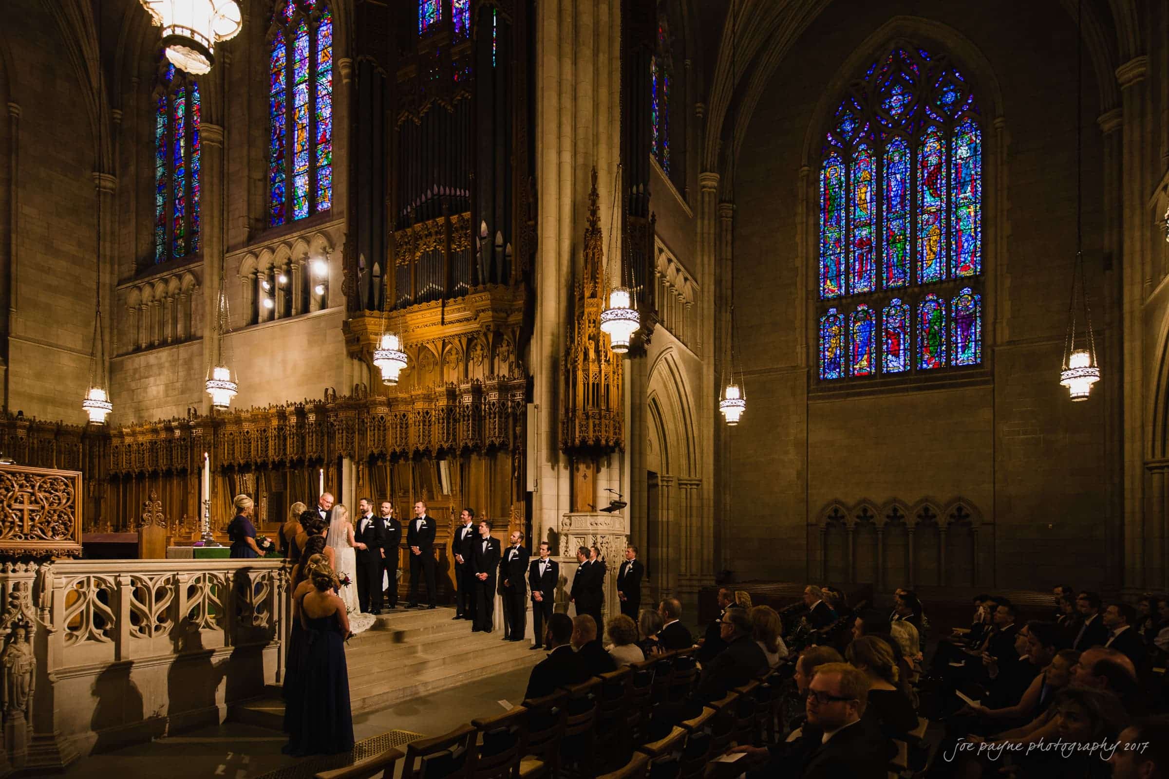 A wedding ceremony inside a grand cathedral. The bride and groom stand at the altar with the bridal party, surrounded by ornate architecture and illuminated by colorful stained glass windows. Guests are seated, observing the ceremony.