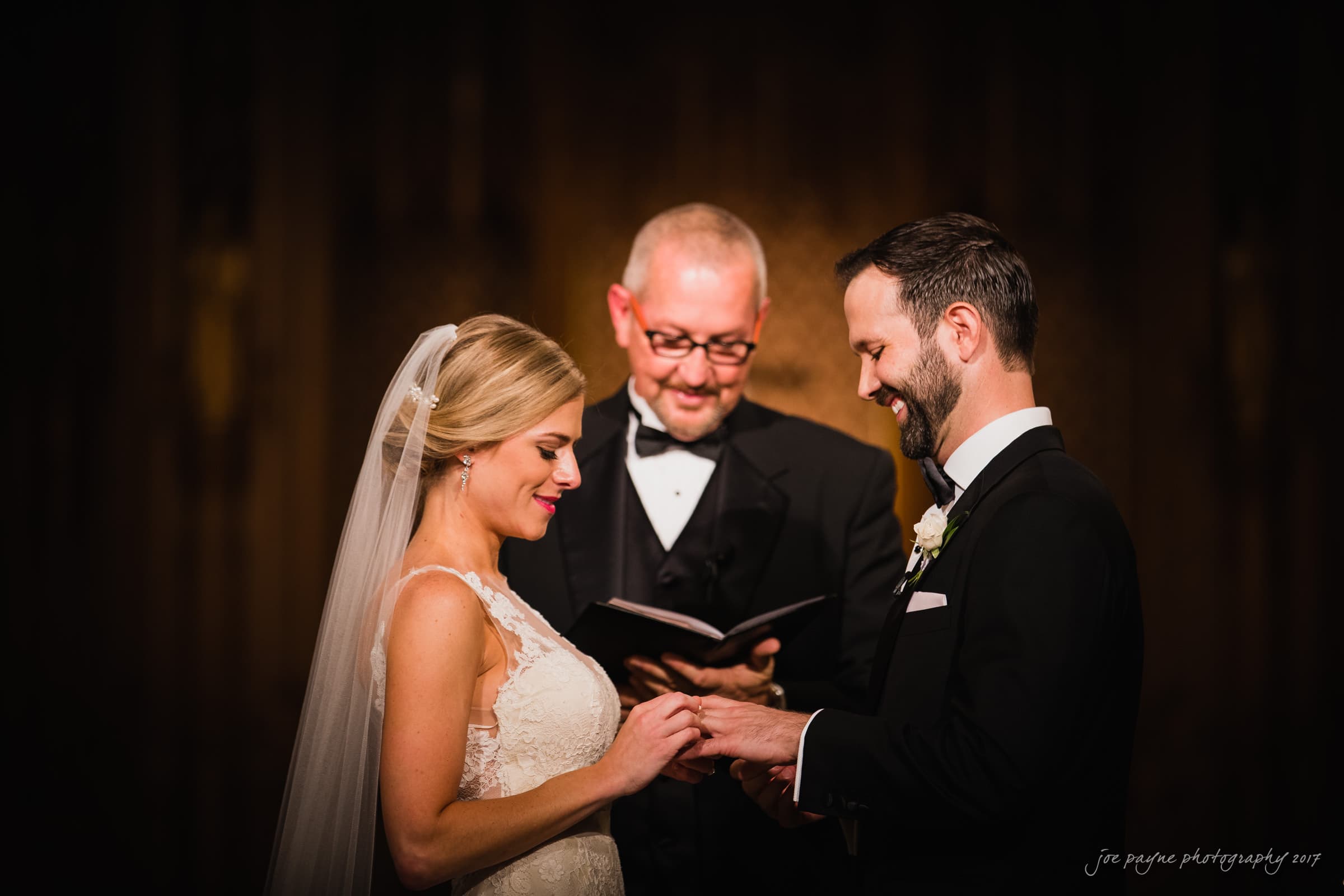 A bride and groom exchange rings during their wedding ceremony. The bride wears a white gown with a veil, and the groom is in a black suit. A man in glasses officiates the ceremony, holding a book and standing in front of a softly lit background.