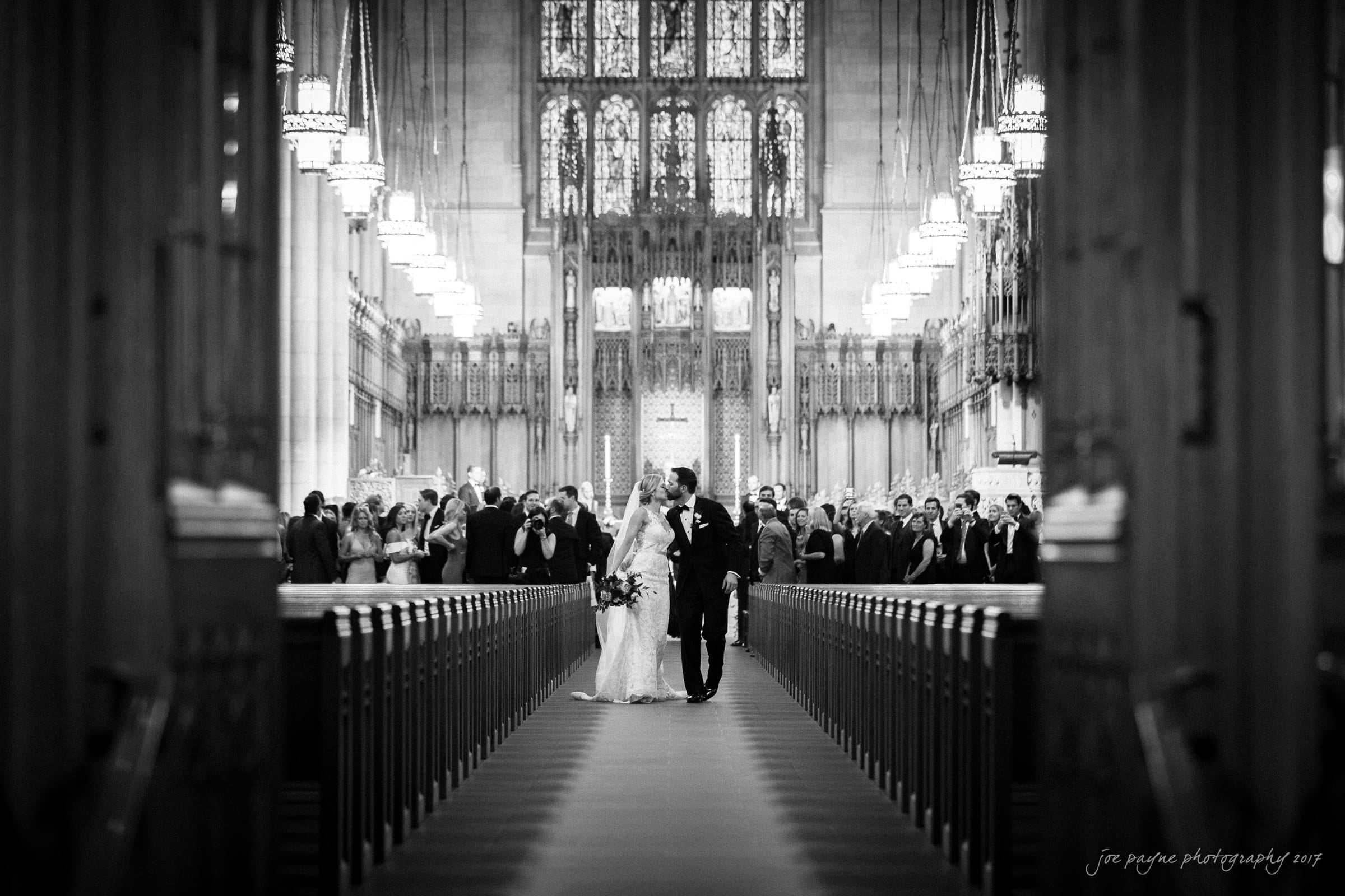 A black and white photo of a bride and groom walking down the aisle of a grand church. The church is filled with guests standing, with high vaulted ceilings and ornate stained glass windows in the background.