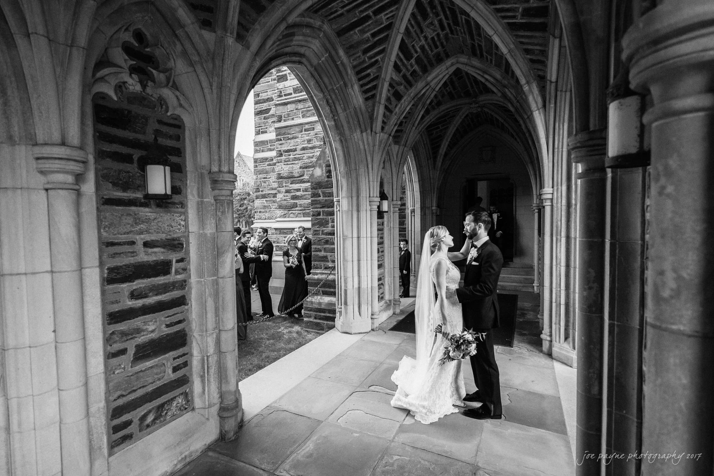 A bride and groom share a kiss under an arched stone walkway. They are surrounded by intricate architecture. In the background, a group of people watches the scene, some taking photos. The atmosphere is elegant and timeless.
