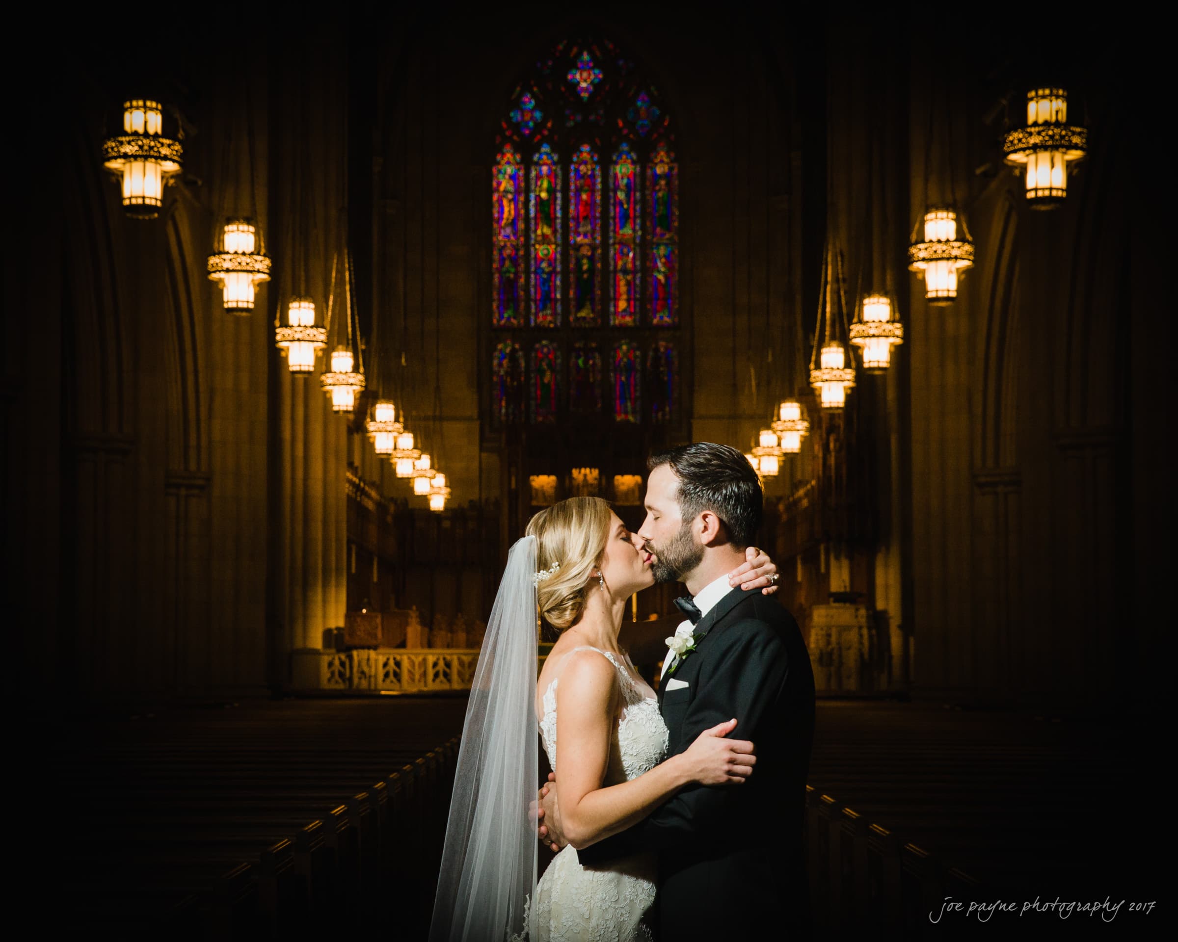 A bride and groom share a kiss in a dimly lit church with ornate stained glass windows and hanging lights. The bride wears a white dress and veil, while the groom is in a black suit. They stand at the center aisle with the altar in the background.