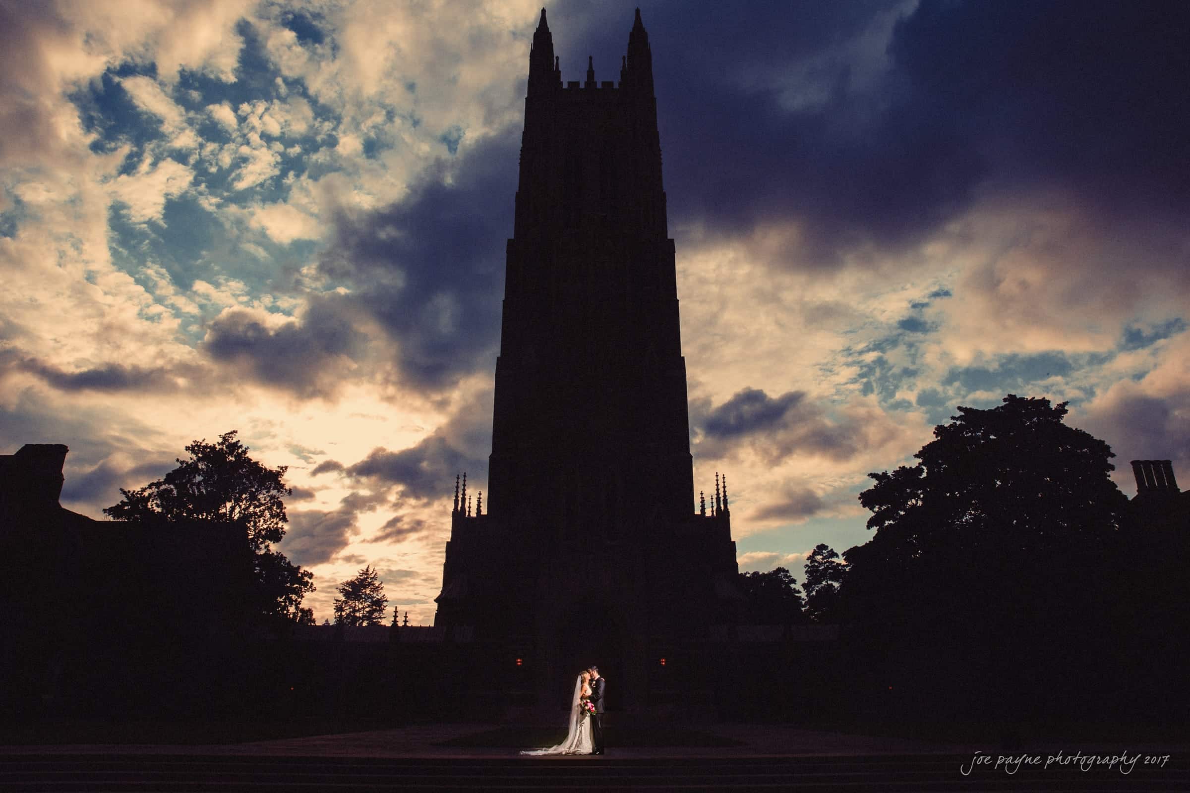 Silhouetted couple stands in front of a tall, dark cathedral at sunset, with dramatic clouds filling the sky. Shadows of trees frame the scene, creating a romantic and atmospheric setting.