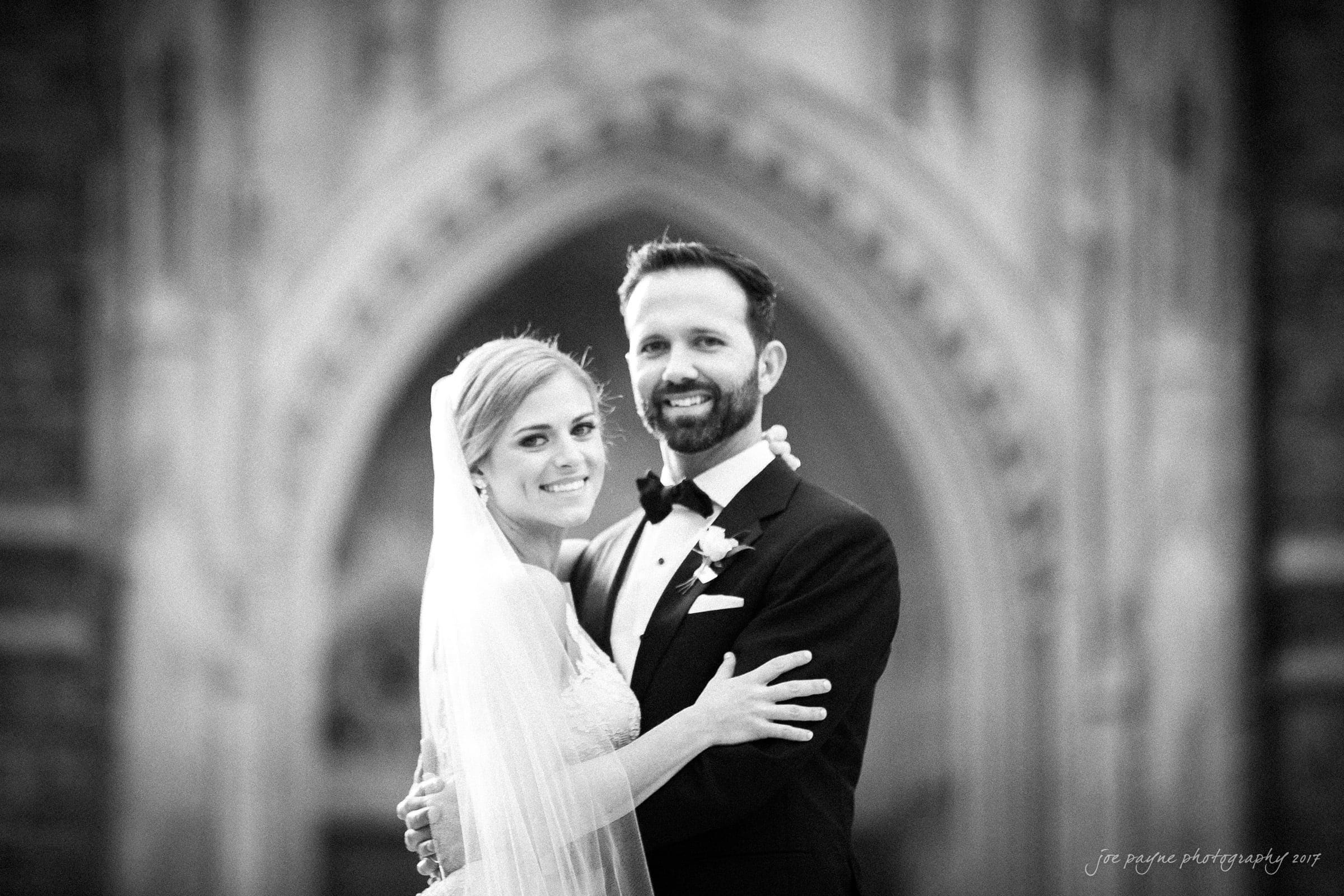 A bride and groom stand in front of an ornate church entrance. The bride is wearing a long veil and a white dress, while the groom is in a black tuxedo. They are smiling and embracing, with the architectural details blurred in the background.