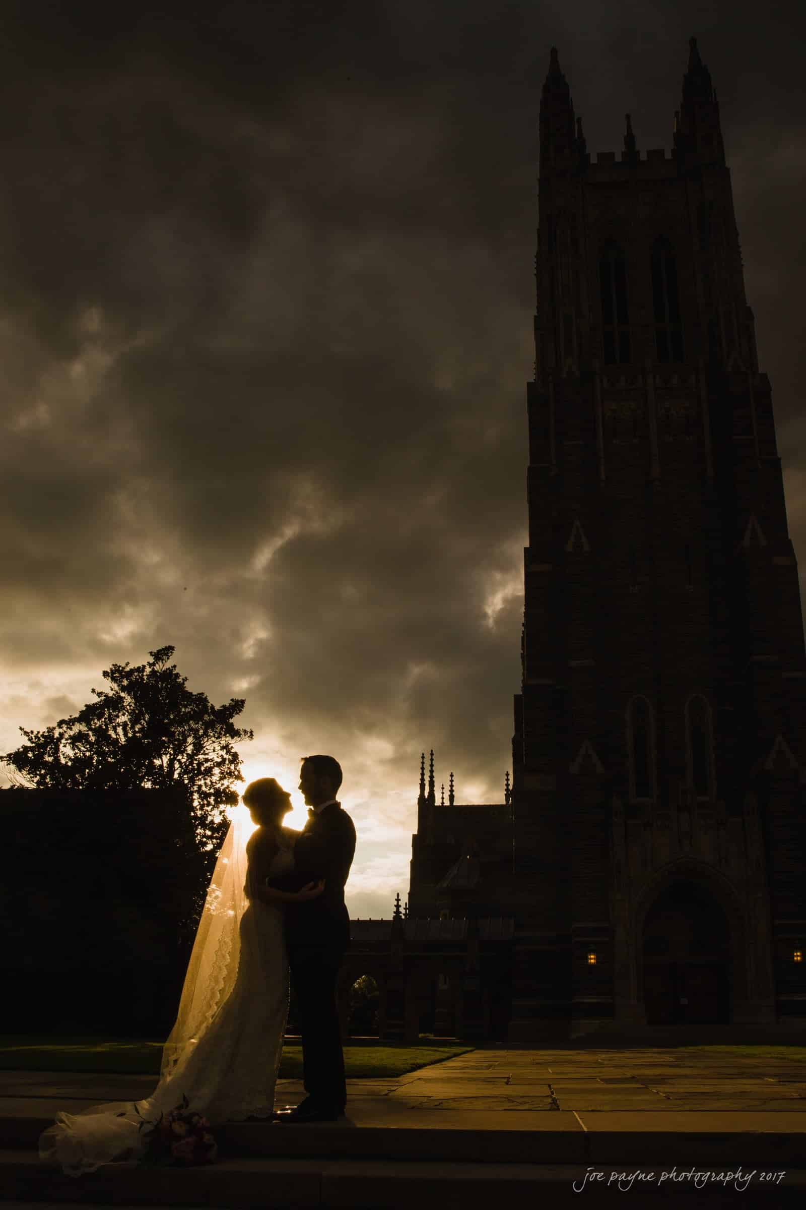 A silhouetted couple embraces in front of a towering cathedral at dusk. The brides veil is gently illuminated by the fading light, and dramatic clouds fill the sky, creating a moody and romantic atmosphere.
