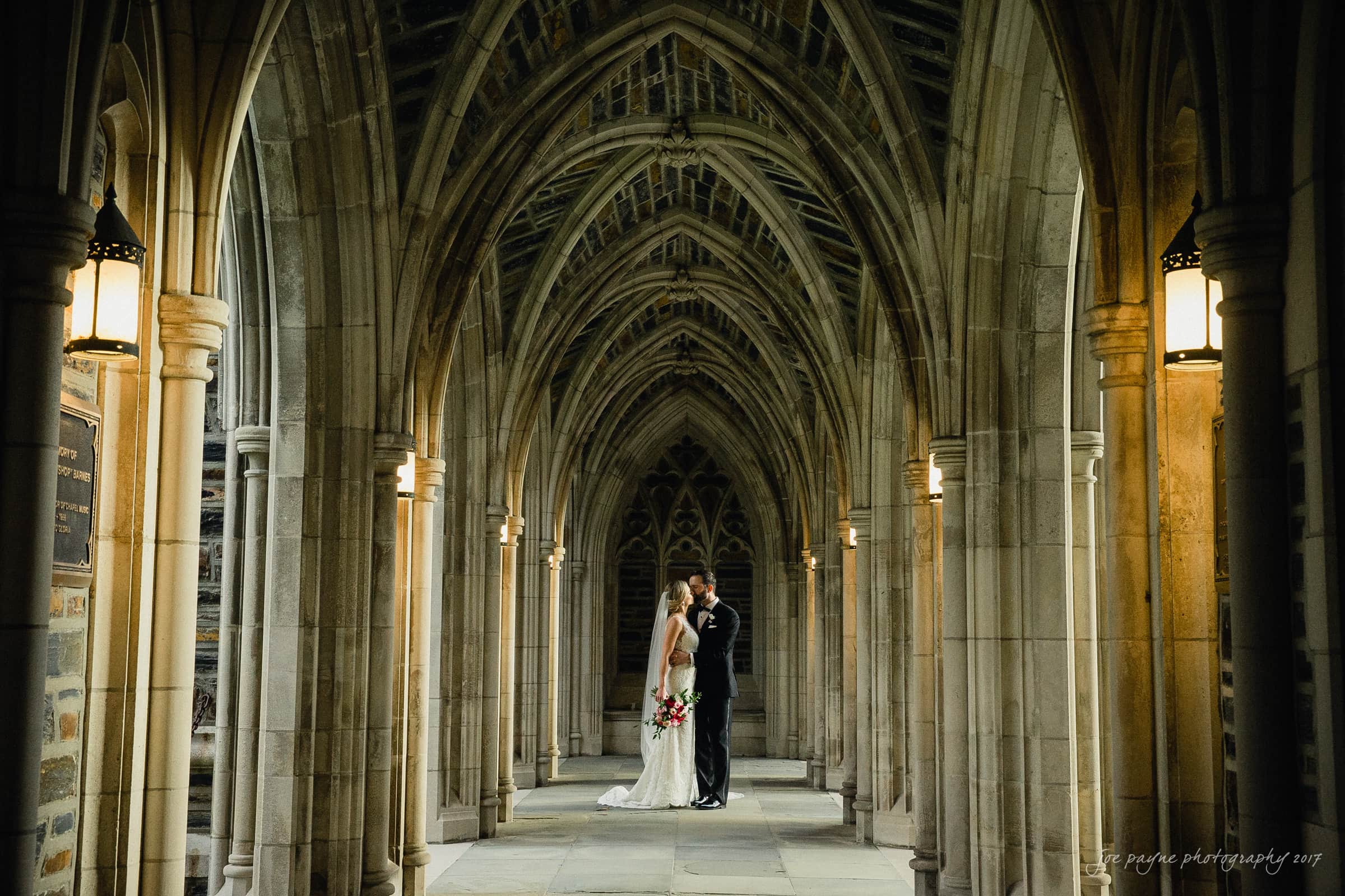 A bride and groom stand embracing in a stone Gothic archway. They are centered under the intricate vaulted ceiling, surrounded by warm light from hanging lanterns. The bride holds a bouquet of red and white flowers.