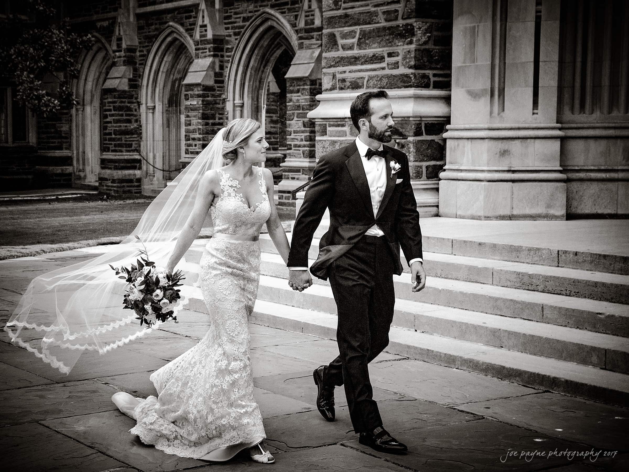 A bride in a lace gown and veil holds hands with a groom in a tuxedo as they walk together outside a stone building with arched doorways. The bride carries a bouquet, and both appear joyful. The scene is in black and white.