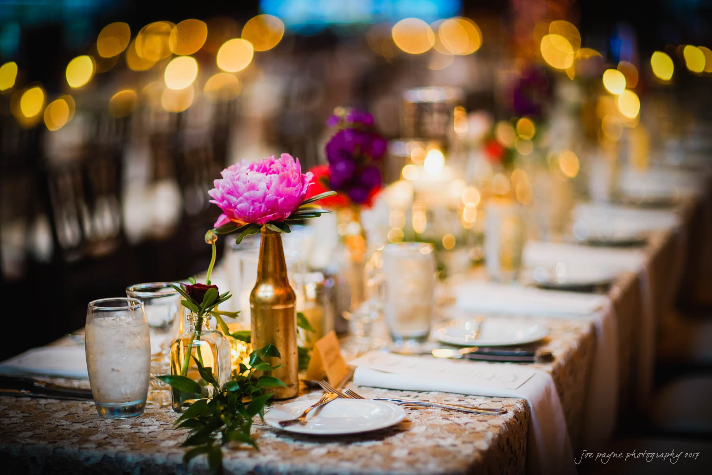 A beautifully set long dining table features floral centerpieces in golden vases and glowing candles. Delicate lace tablecloths, neatly arranged glassware, cutlery, and folded napkins create an elegant and inviting ambiance.
