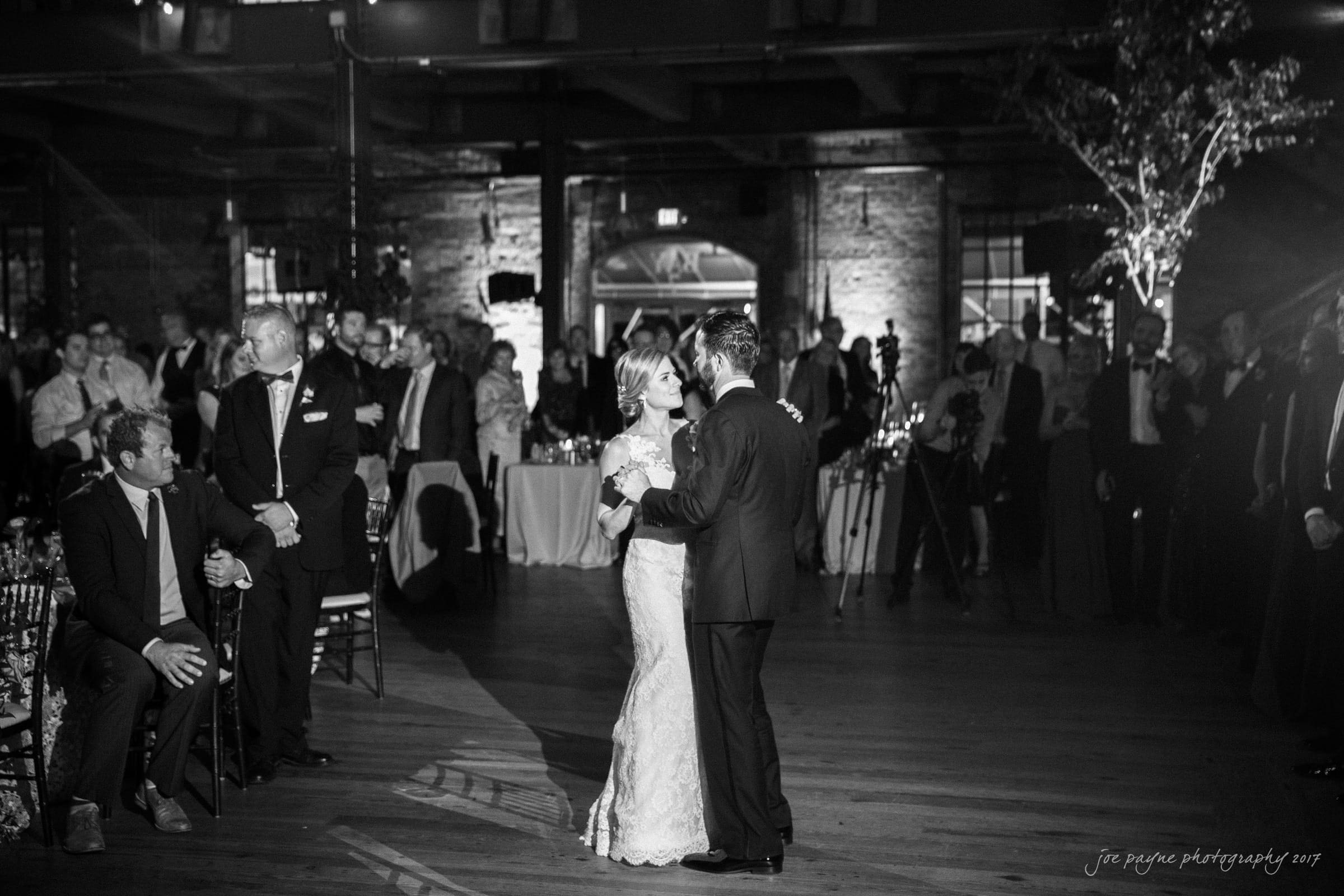 A bride and groom share their first dance in a dimly lit, elegant venue. Guests watch from their seats, with some standing around the wooden dance floor. A large archway and decorative lighting enhance the romantic atmosphere.