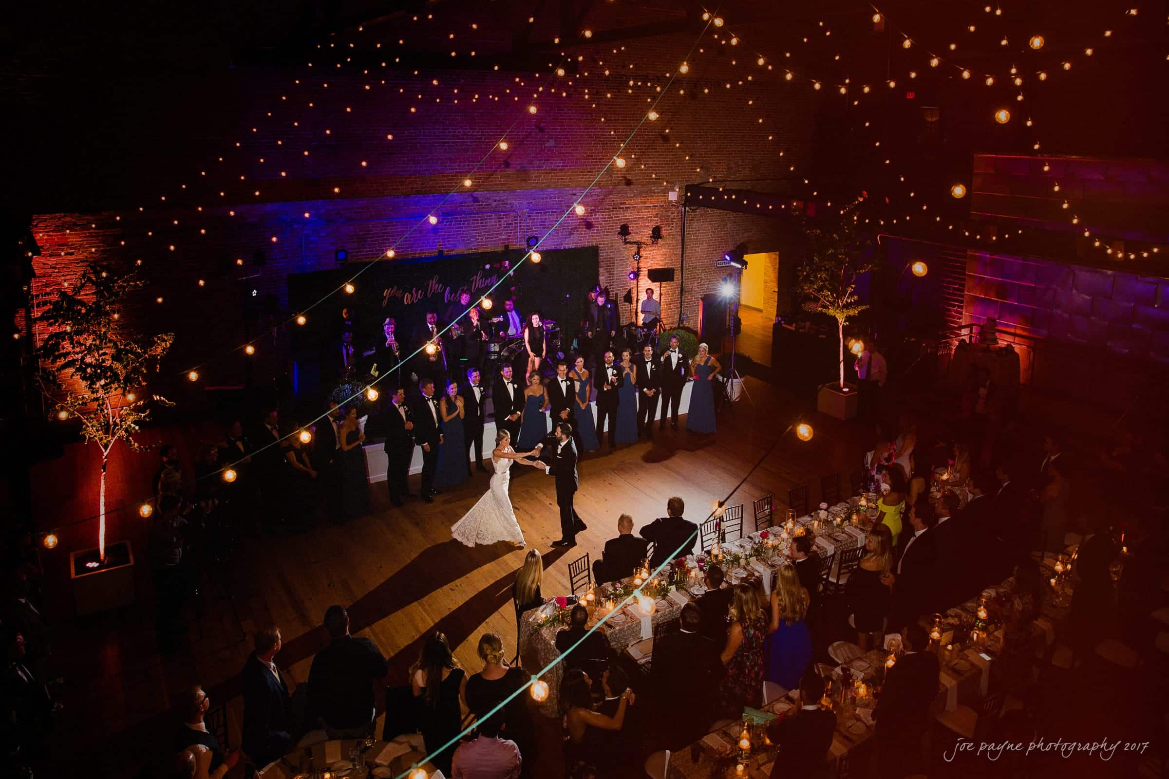 A bride and groom dance under string lights in a warmly lit venue. Guests stand around the perimeter watching, with tables elegantly set for a wedding reception.