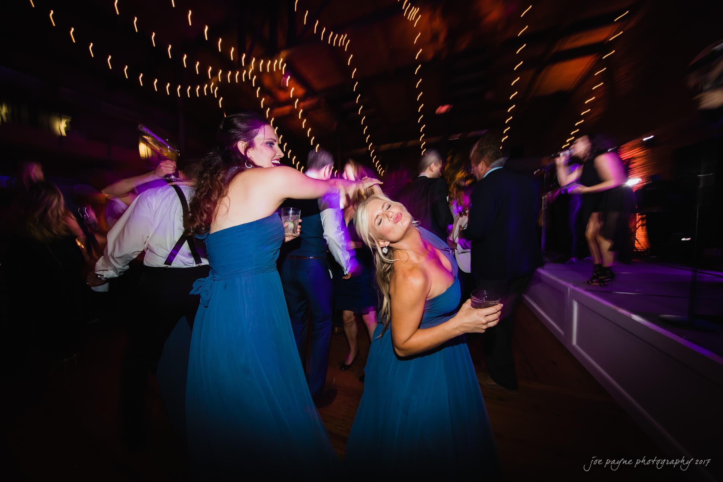 People dancing energetically at a party, with two women in matching blue dresses in the foreground. Warm string lights hang above, and a band performs on a stage to the right. The ambiance is lively and festive.