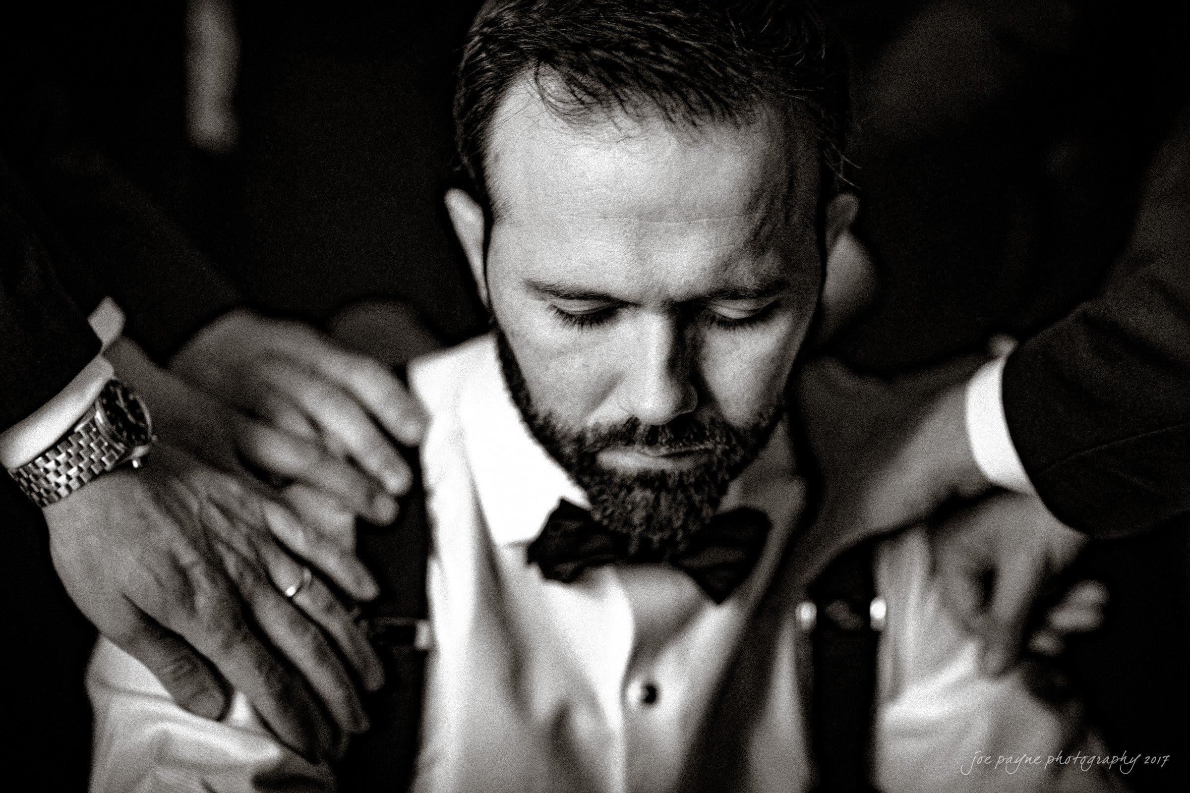 A man with a beard and bow tie is sitting with his head down, while several hands rest on his shoulders in a gesture of support and solidarity. The image is in black and white, highlighting an emotional and intimate moment.