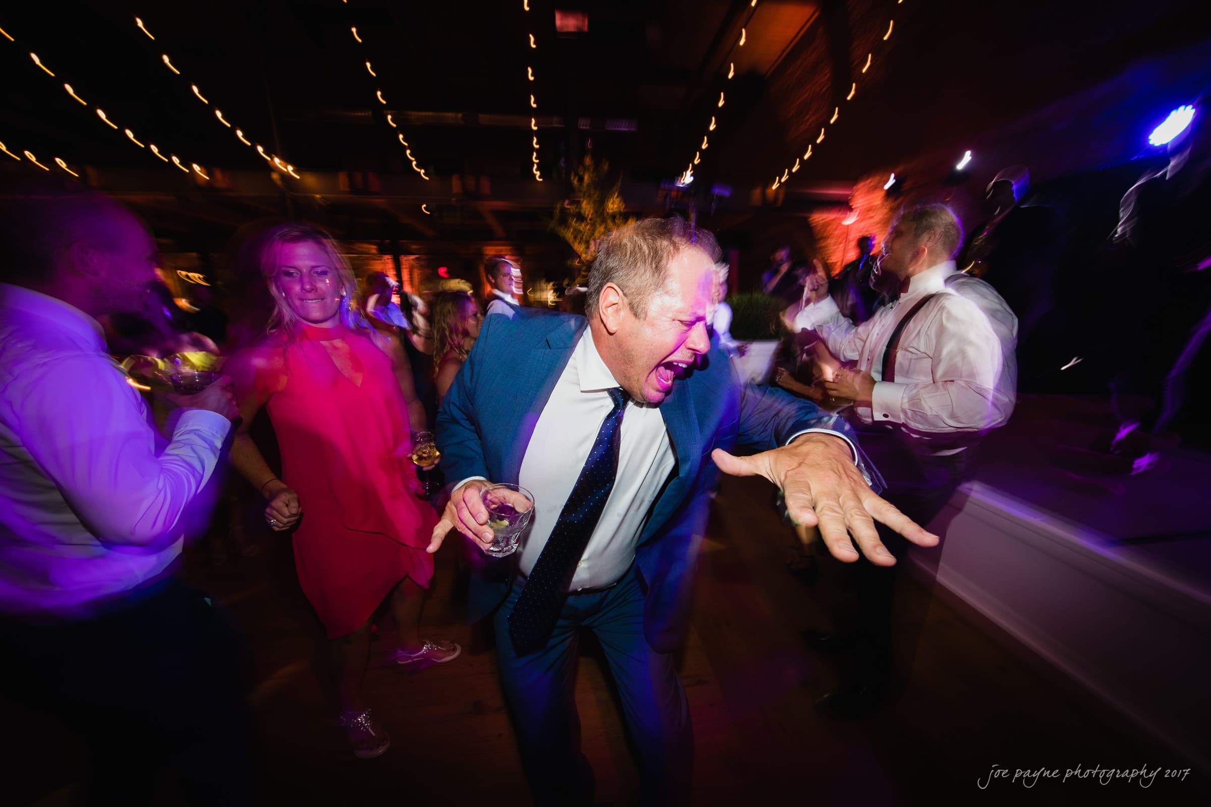 A man in a blue suit energetically dances at a party with blurred lights and other guests in the background. The lively atmosphere is highlighted by colorful lighting and motion.