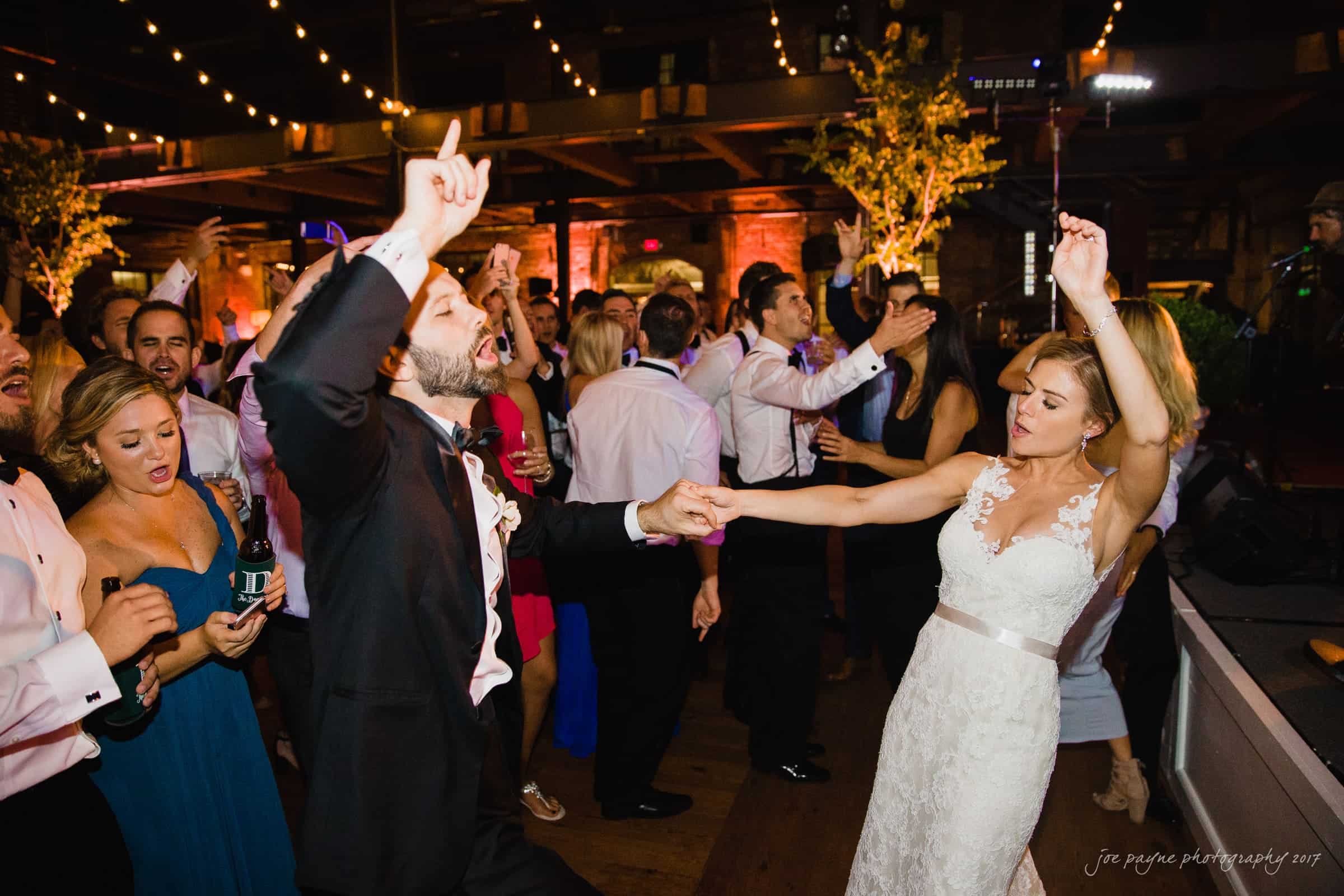 A joyful bride and groom dance energetically on a lively dance floor at a wedding reception. They are surrounded by friends and guests, with string lights illuminating the venue, creating a festive and celebratory atmosphere.