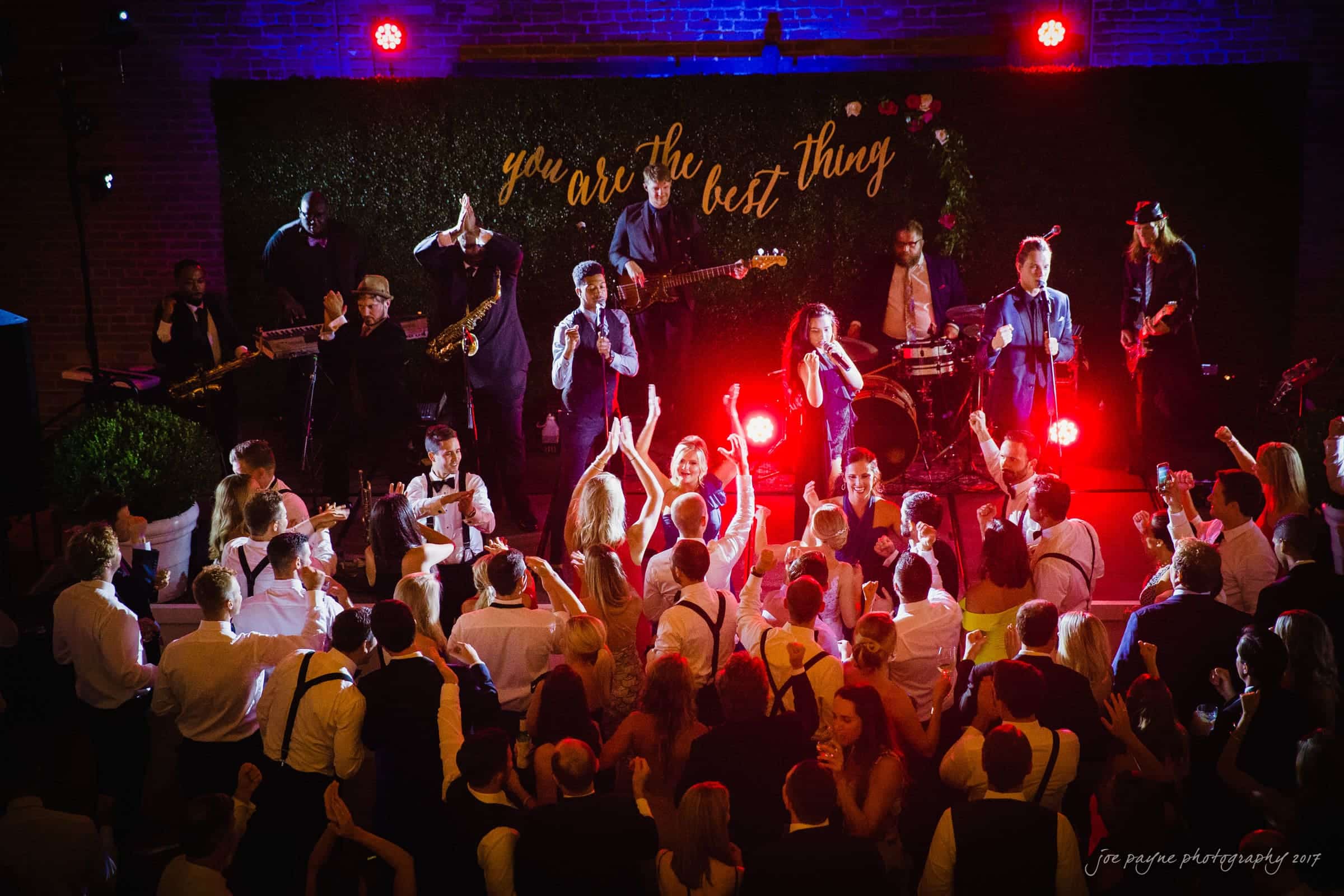 A lively wedding reception with a band performing on stage. Guests are dancing energetically on the dance floor. A neon sign reading you are the best thing is illuminated in the background.