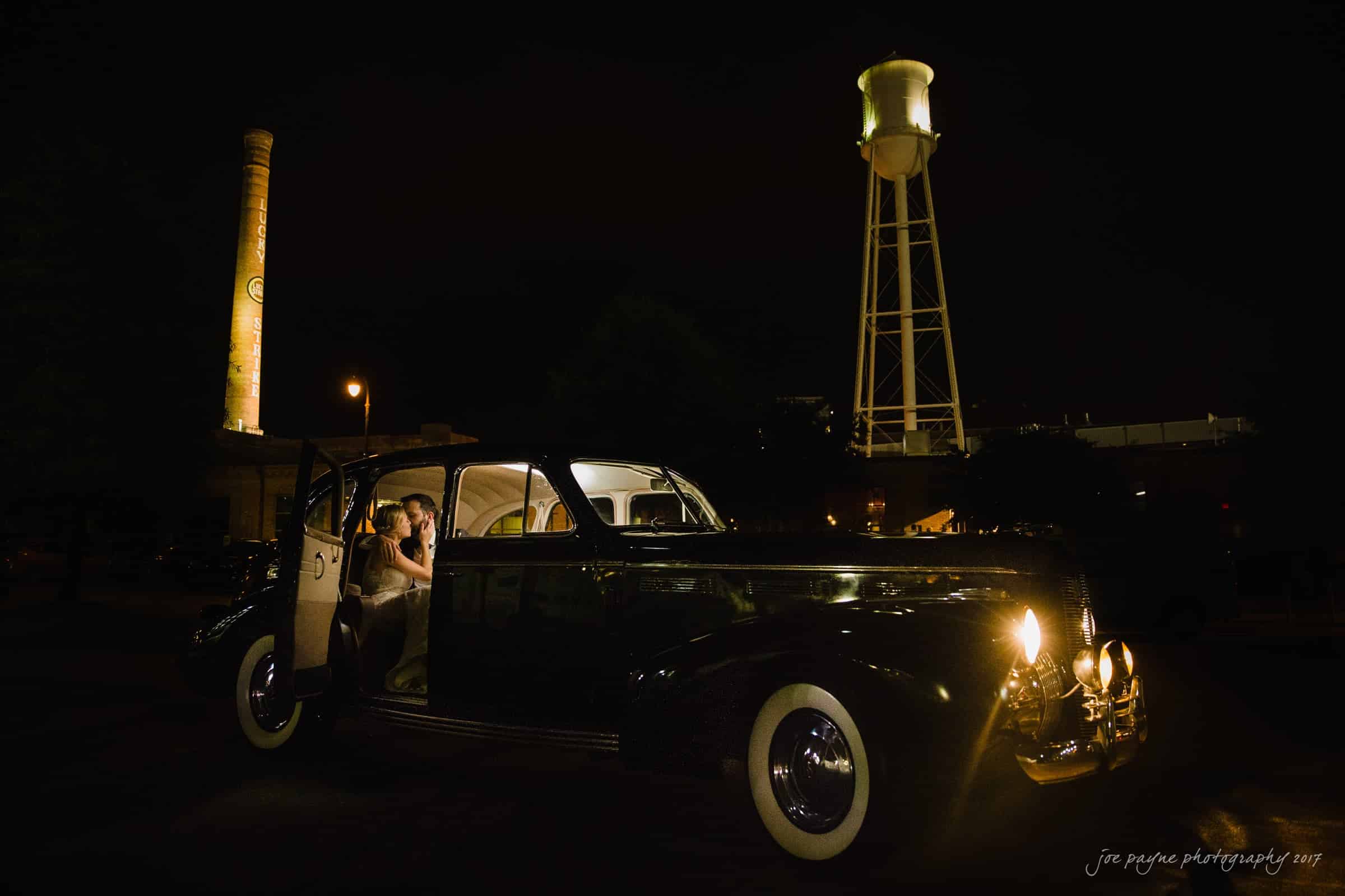 A vintage car is parked at night under a brightly lit water tower. A person sits inside the car with the door open, illuminated by the cars interior light. An industrial chimney is visible in the background.