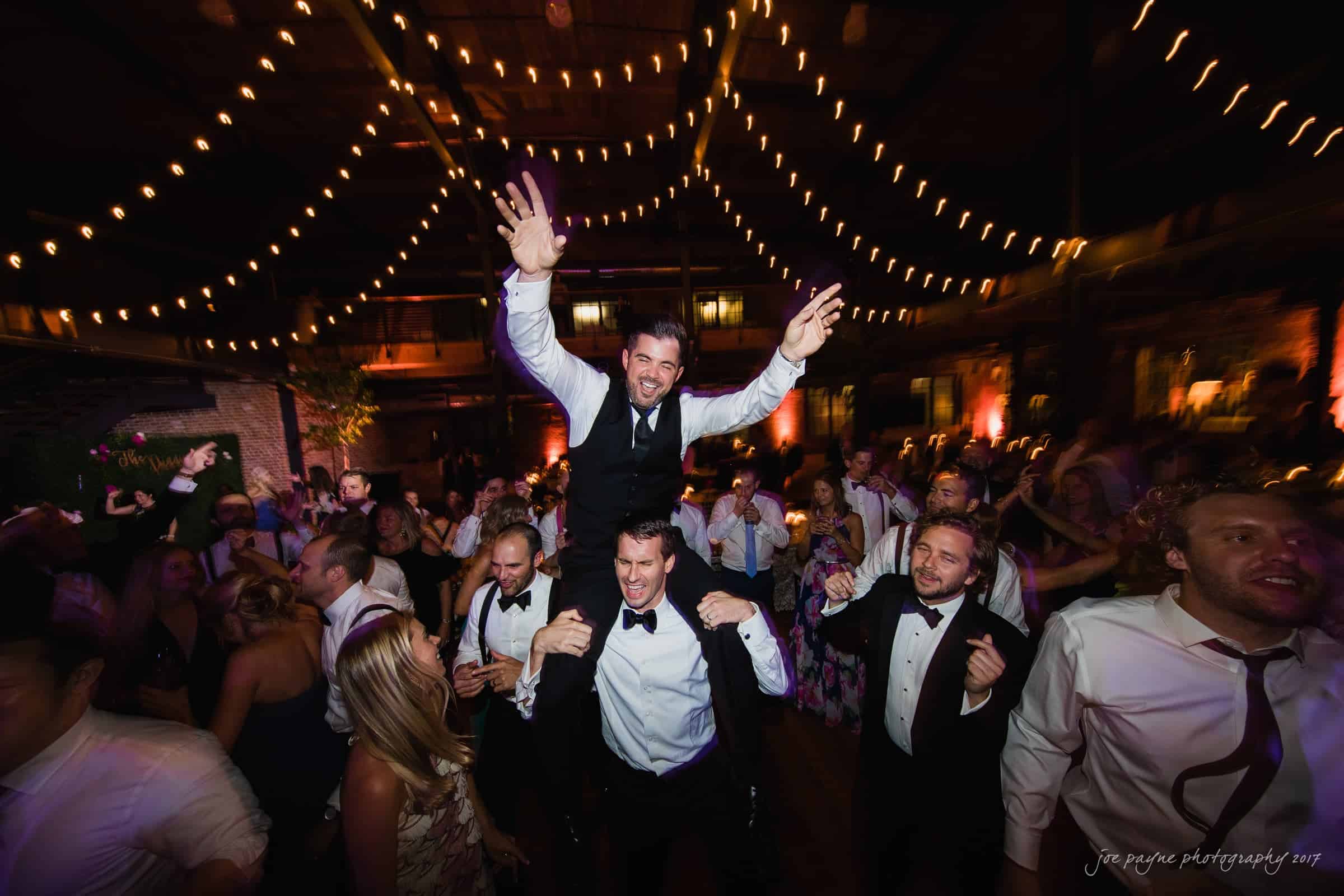 A lively wedding celebration with guests dancing under string lights. A man is joyfully lifted onto another guest’s shoulders, wearing a suit and smiling. The festive atmosphere is highlighted by the warm lights and cheering crowd.