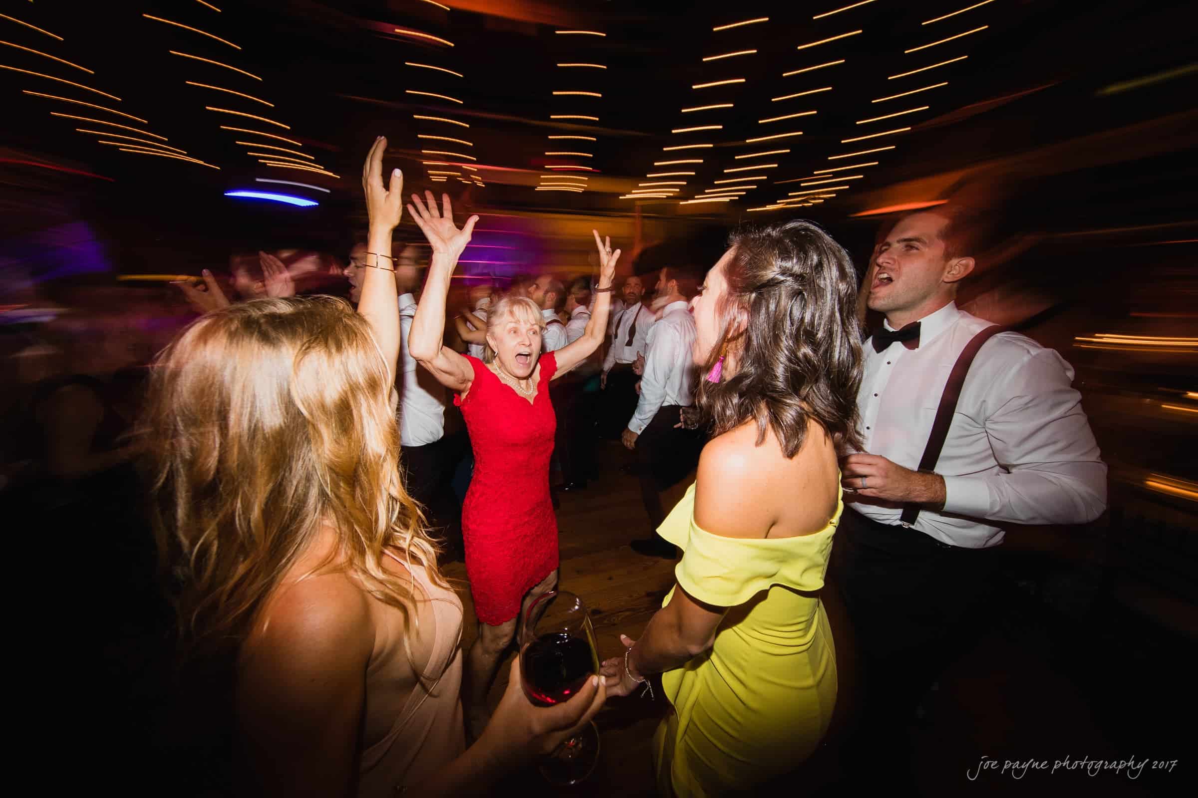 A lively dance floor scene with people dressed formally. A woman in a red dress joyfully raises her arms, while others around her dance and converse. The image has a motion blur and warm, festive lighting.