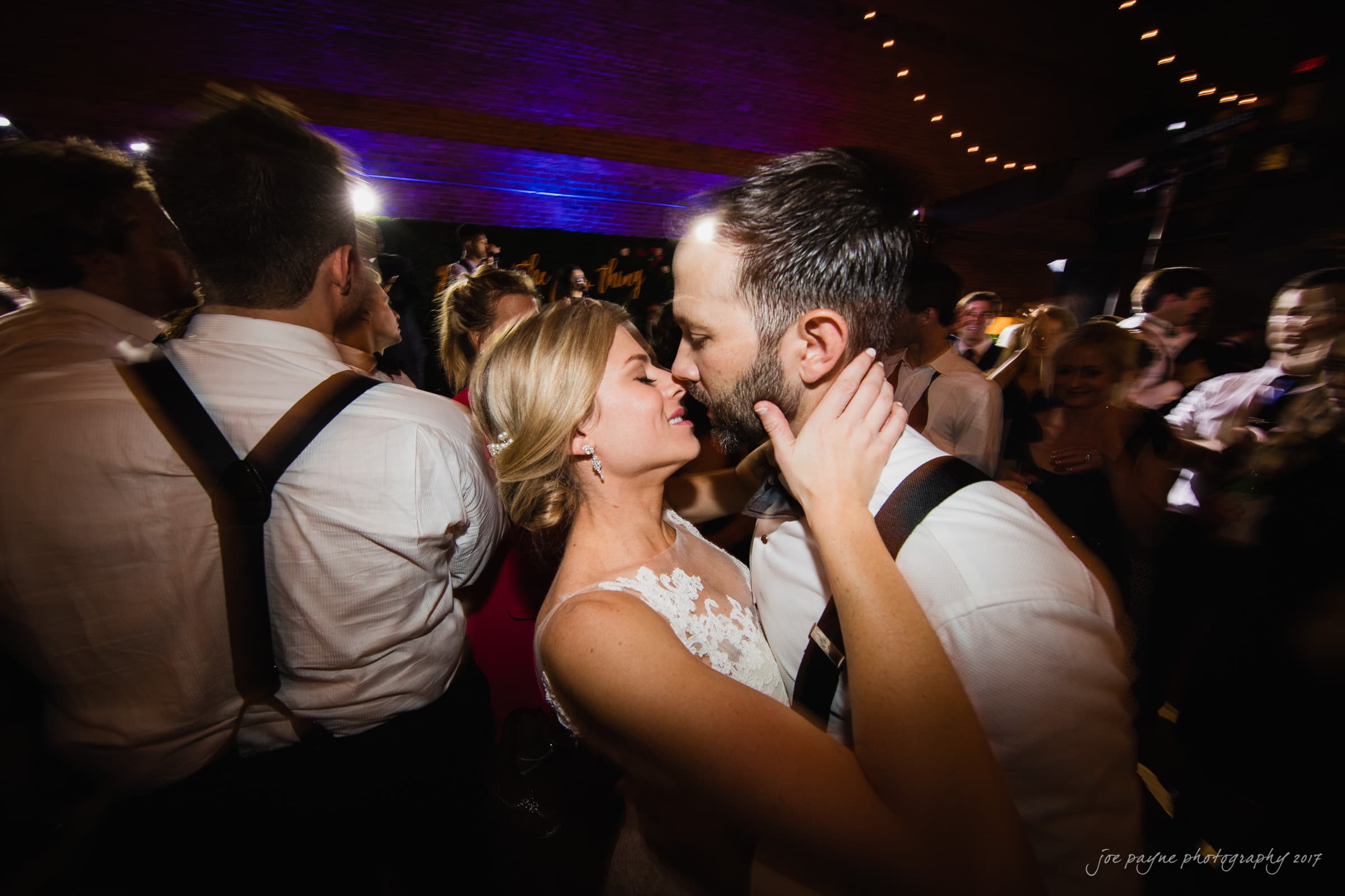 A bride and groom dance closely, surrounded by guests. The bride wears a lace wedding dress, and the groom is in a white shirt with suspenders. Strings of lights create a festive atmosphere as they embrace on the dance floor.