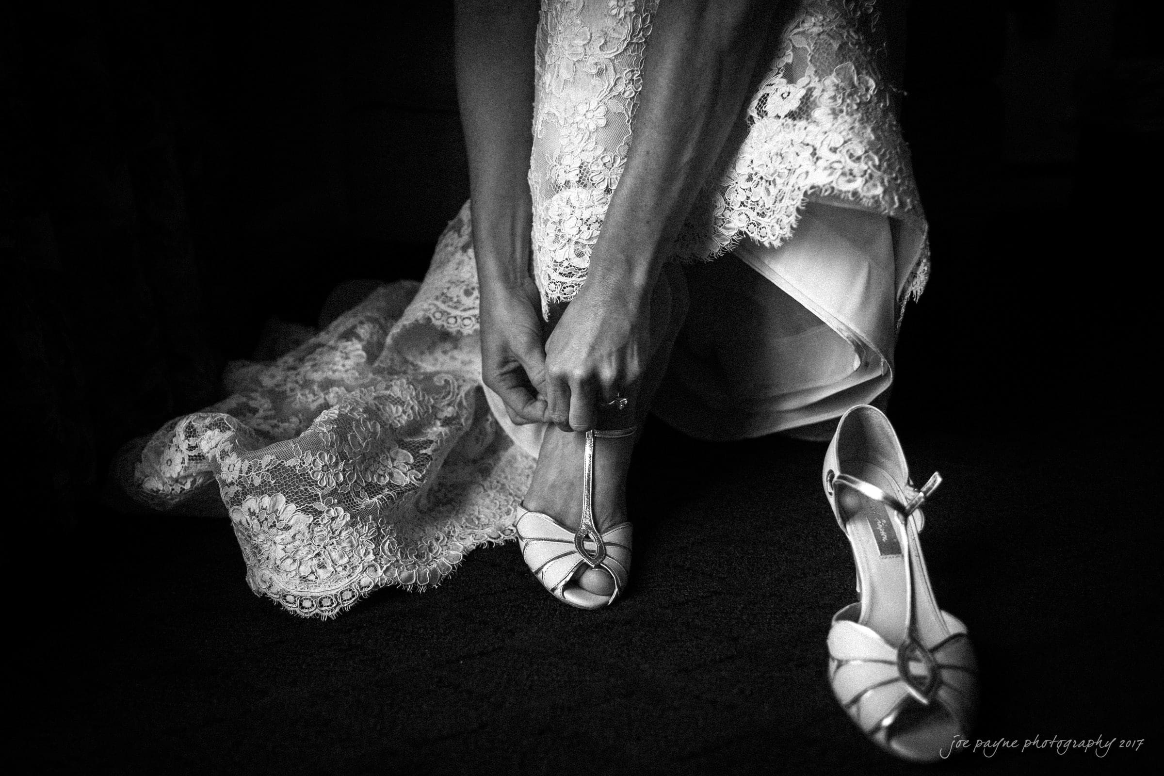 A bride in a lace gown kneels, adjusting the strap of one white high-heeled sandal. The other shoe is on the floor beside her. The image is in black and white, capturing an intimate, candid moment.