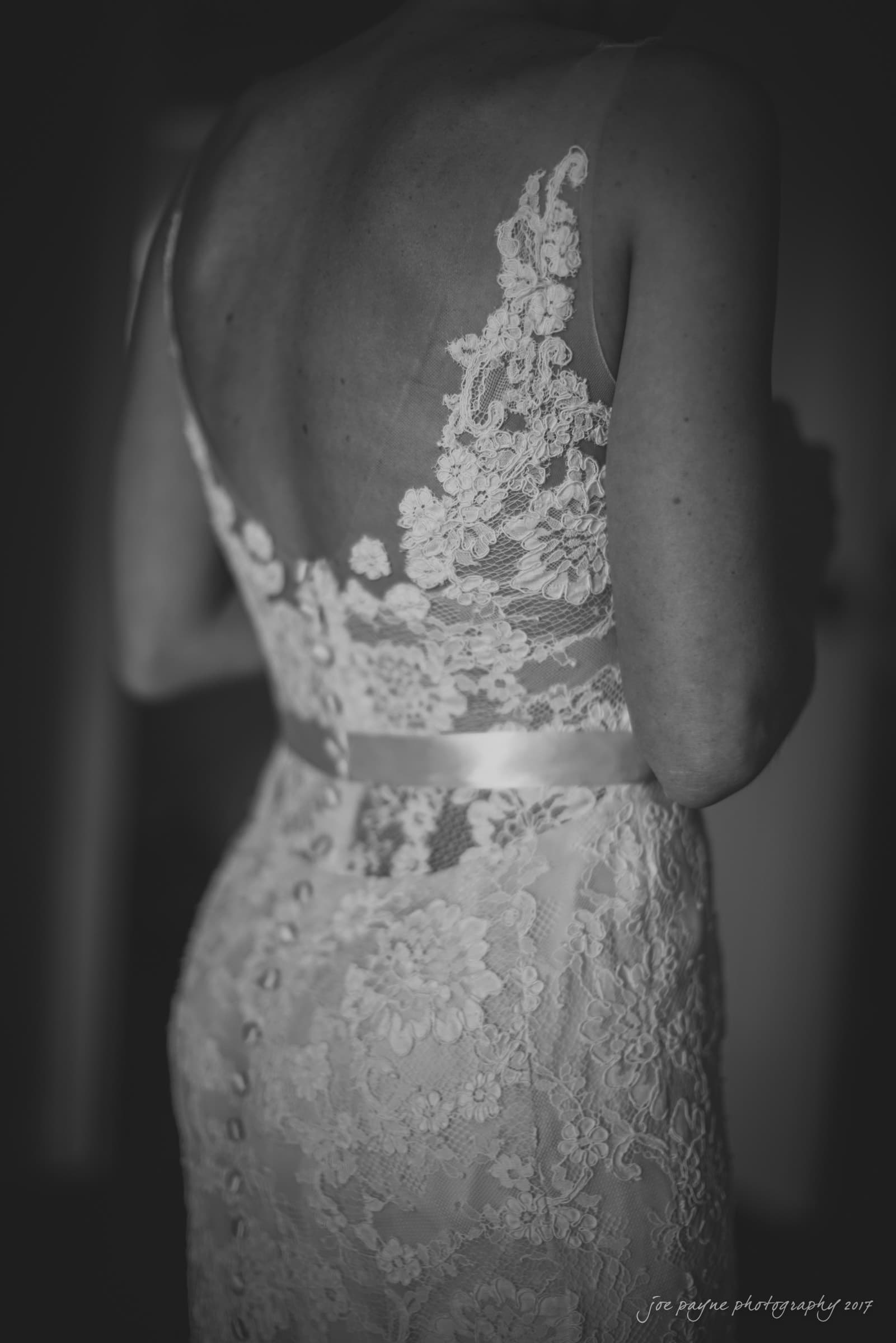 A woman in a detailed lace wedding gown with intricate floral patterns is shown from behind. The dress features a fitted design and buttons down the back, highlighted in soft, monochrome lighting.