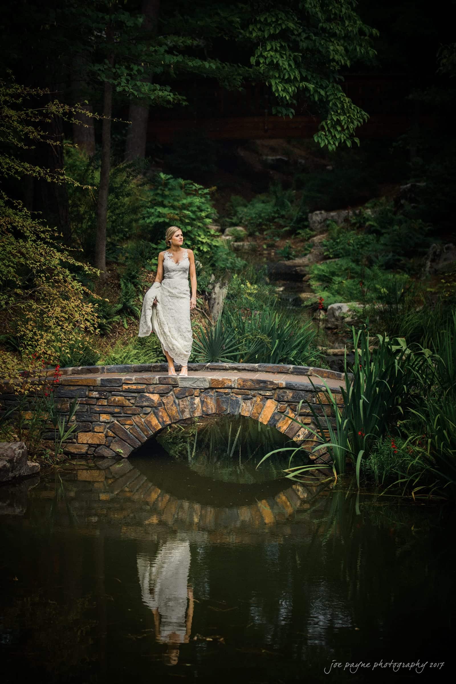 A woman in a white dress stands barefoot on a stone bridge over a pond surrounded by lush greenery and trees. She holds her shoes in one hand, gazing peacefully into the distance.