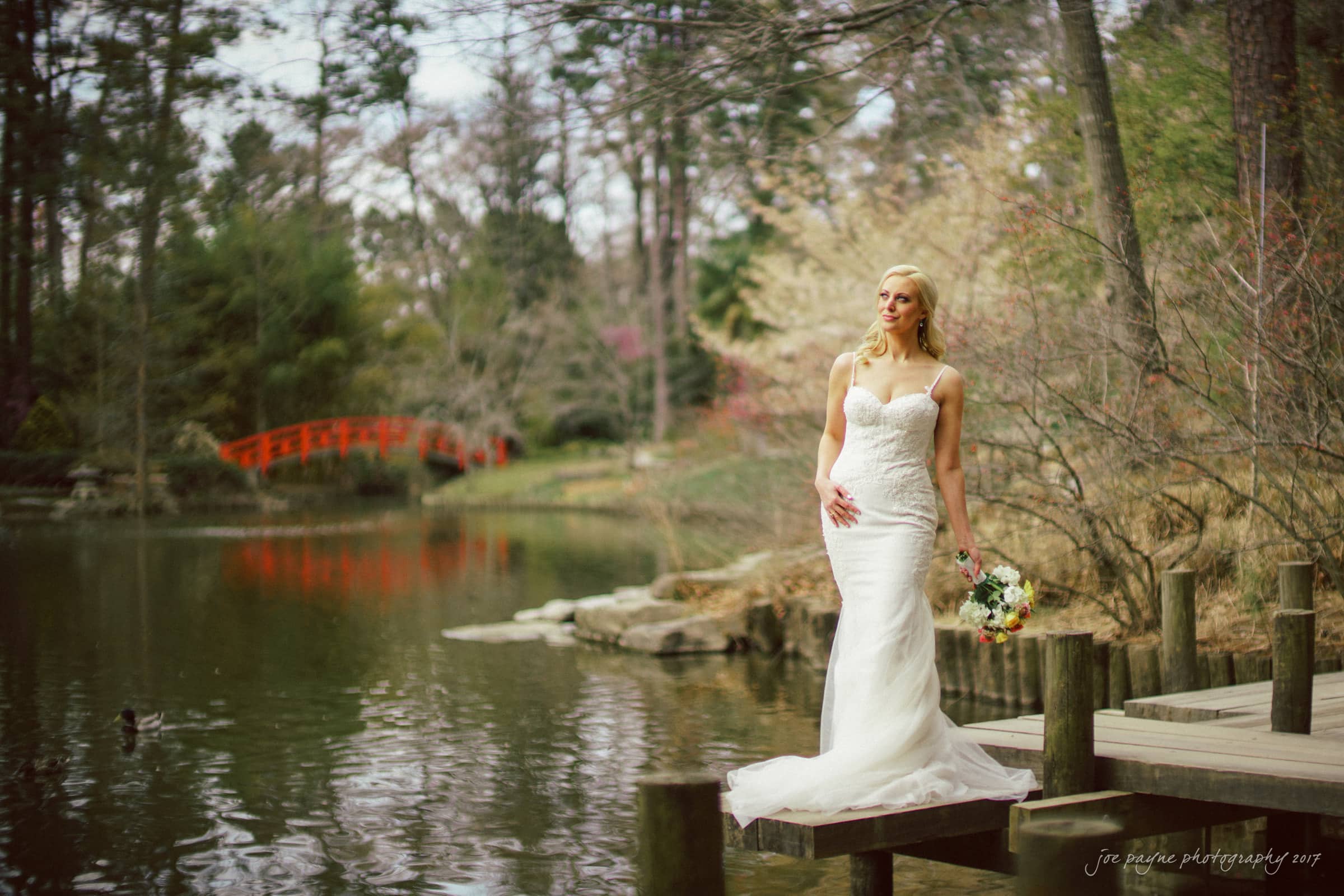A bride in a white wedding dress stands on a wooden dock by a pond, holding a bouquet. Trees and a red footbridge add charm to the scene—serene and surrounded by nature, perfect for duke gardens wedding photography at elizabeth's bridal session.
