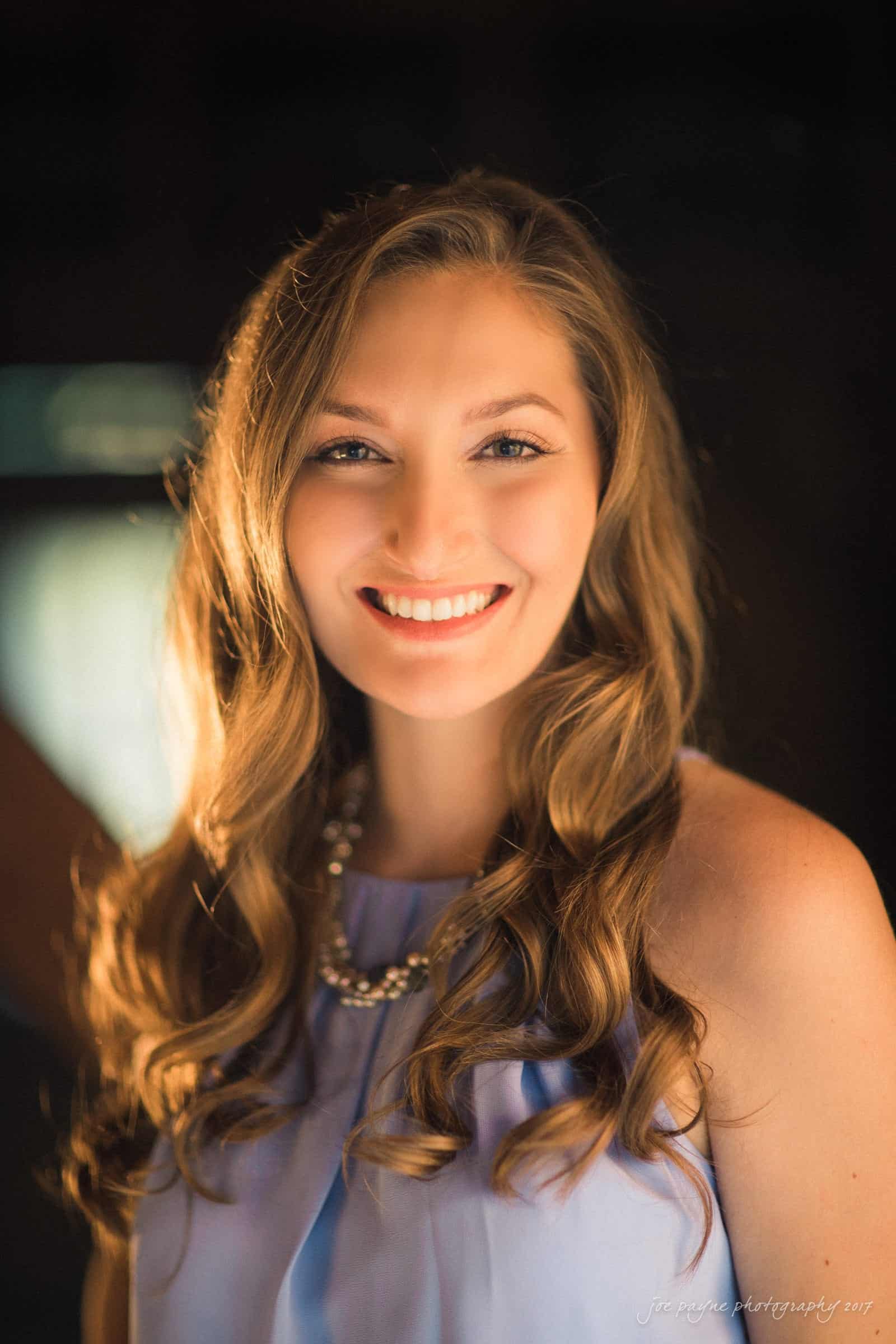 A woman with long, wavy hair smiles warmly. She wears a sleeveless lavender top and a pearl necklace. The background is softly blurred with warm lighting.