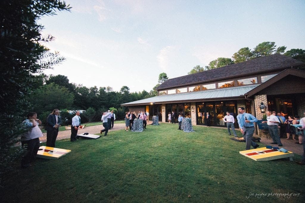 People are gathered on a lawn outside a rustic building during a social event. Some are playing cornhole, while others stand and mingle. The scene is set against a backdrop of trees and a soft evening sky.