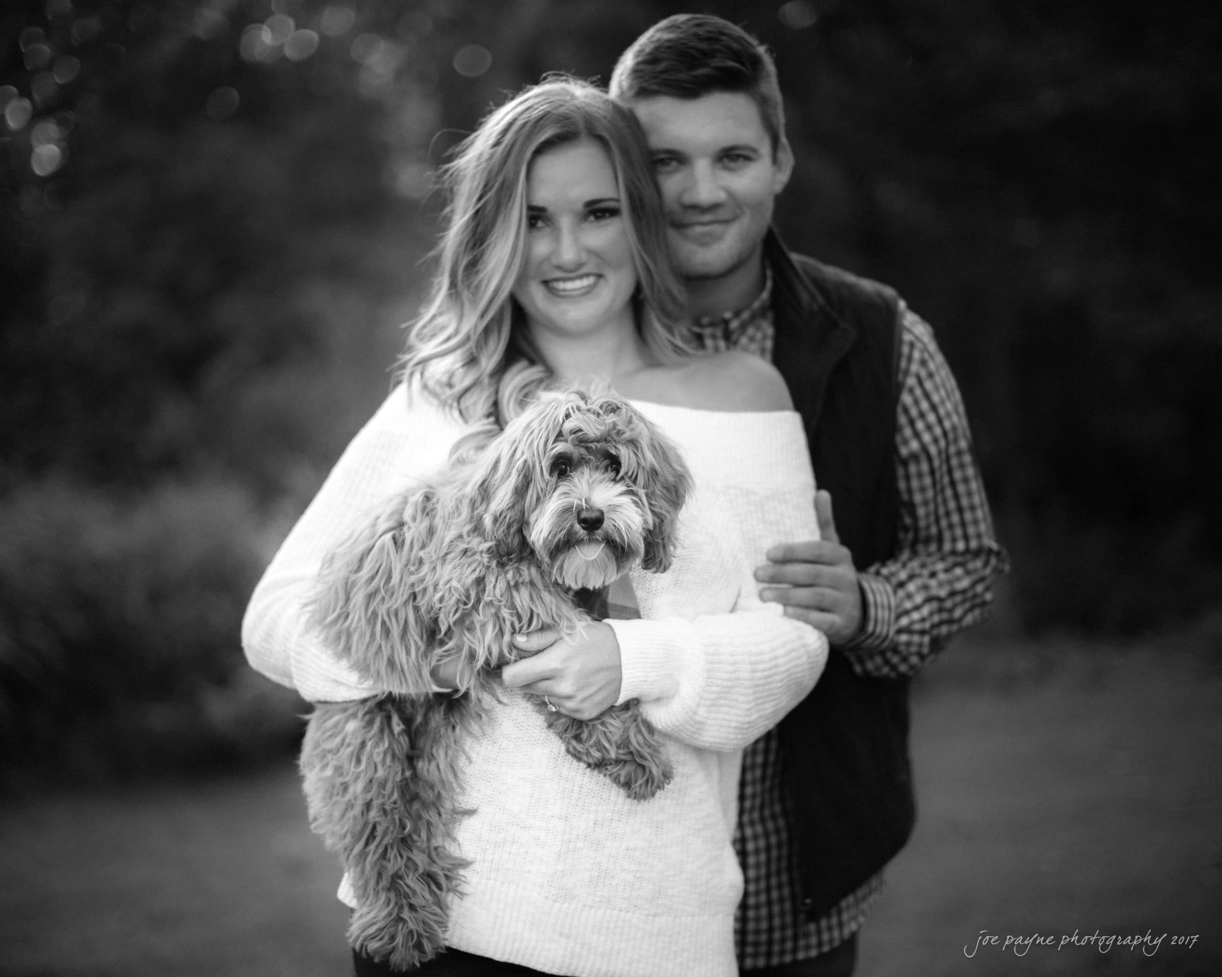 A smiling woman holds a fluffy dog in front of her, while a man stands behind with his hand on her shoulder. Both are outdoors with a blurred natural background. The image is in black and white.