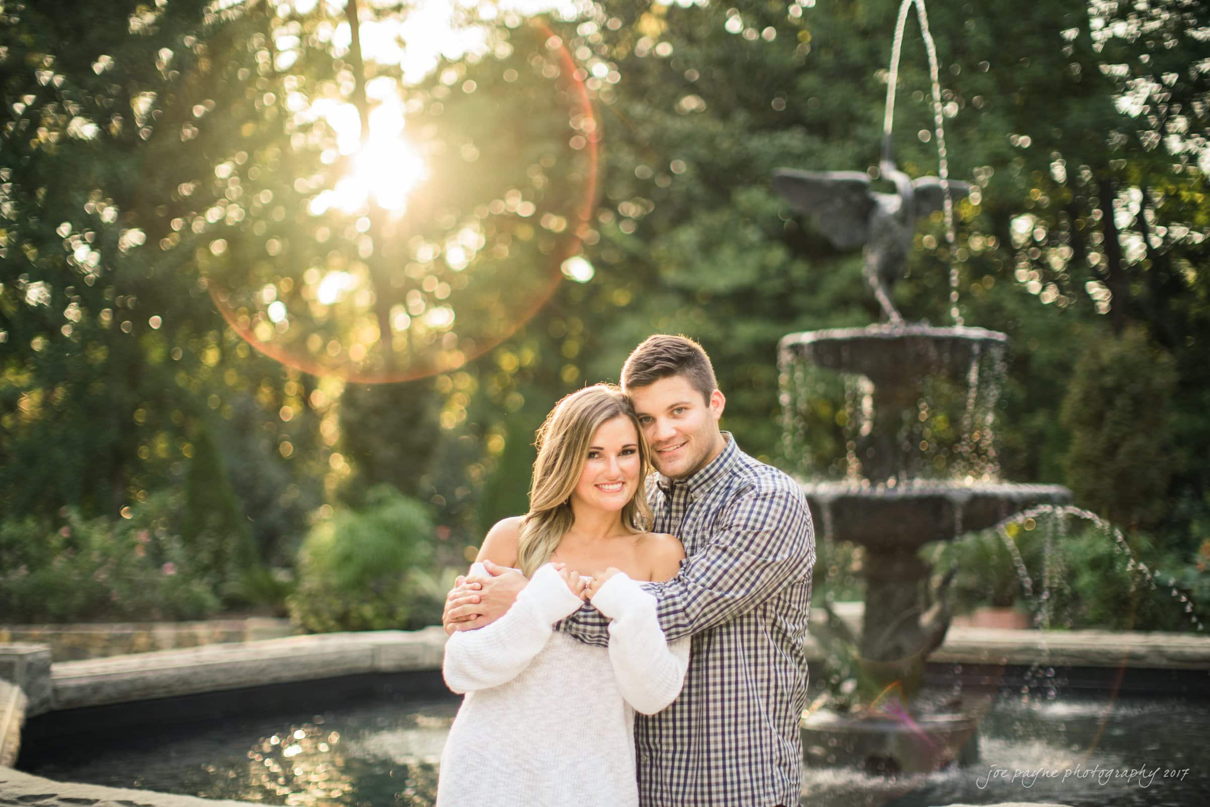 A couple embraces in front of a flowing fountain with an angel statue on top. The sun shines through trees in the background, casting a warm glow around them. The man wears a plaid shirt and the woman a white off-shoulder sweater.