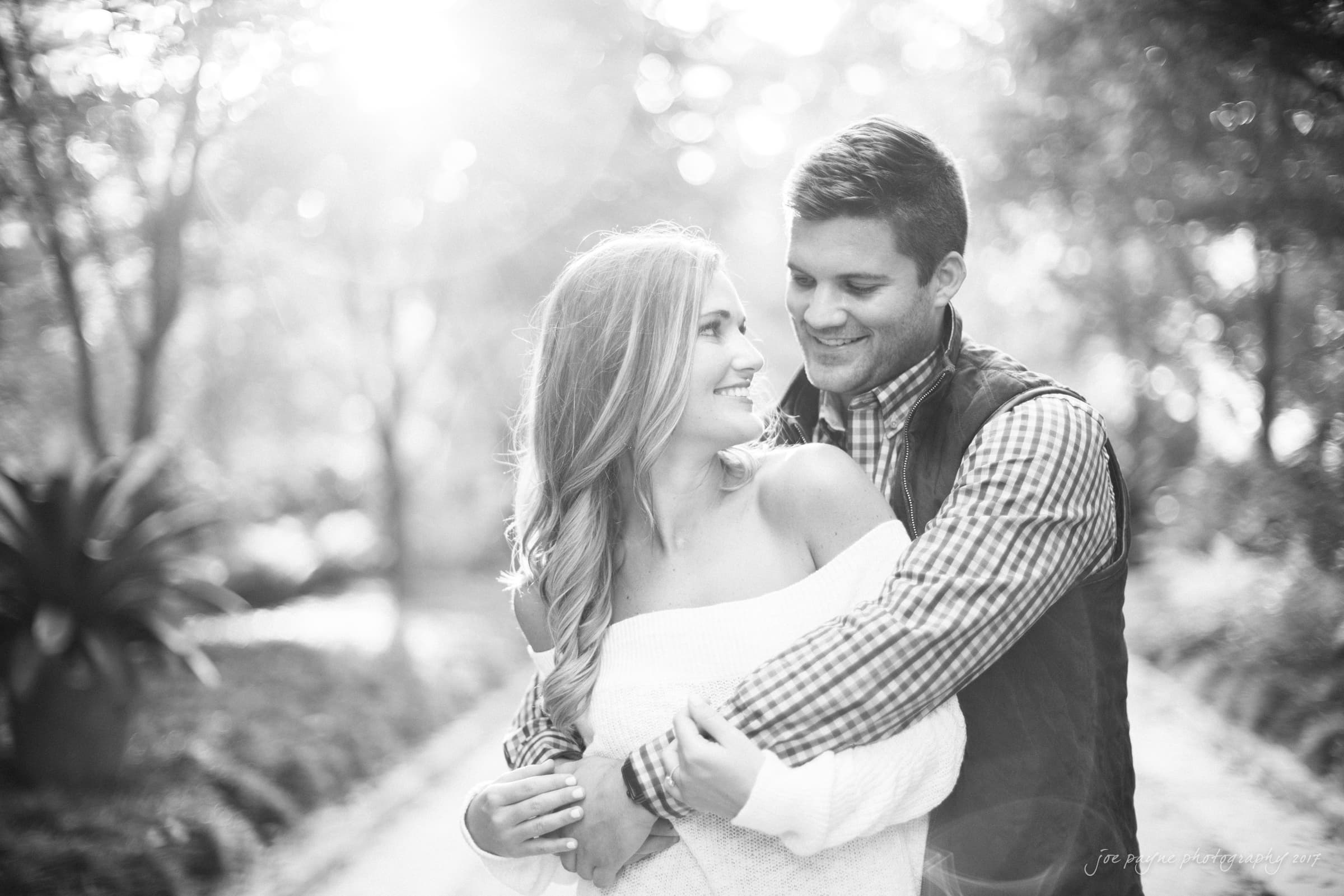 A black and white photo of a couple embracing. The woman is looking at the man with a smile, and they are surrounded by soft foliage and gentle sunlight filtering through the trees.