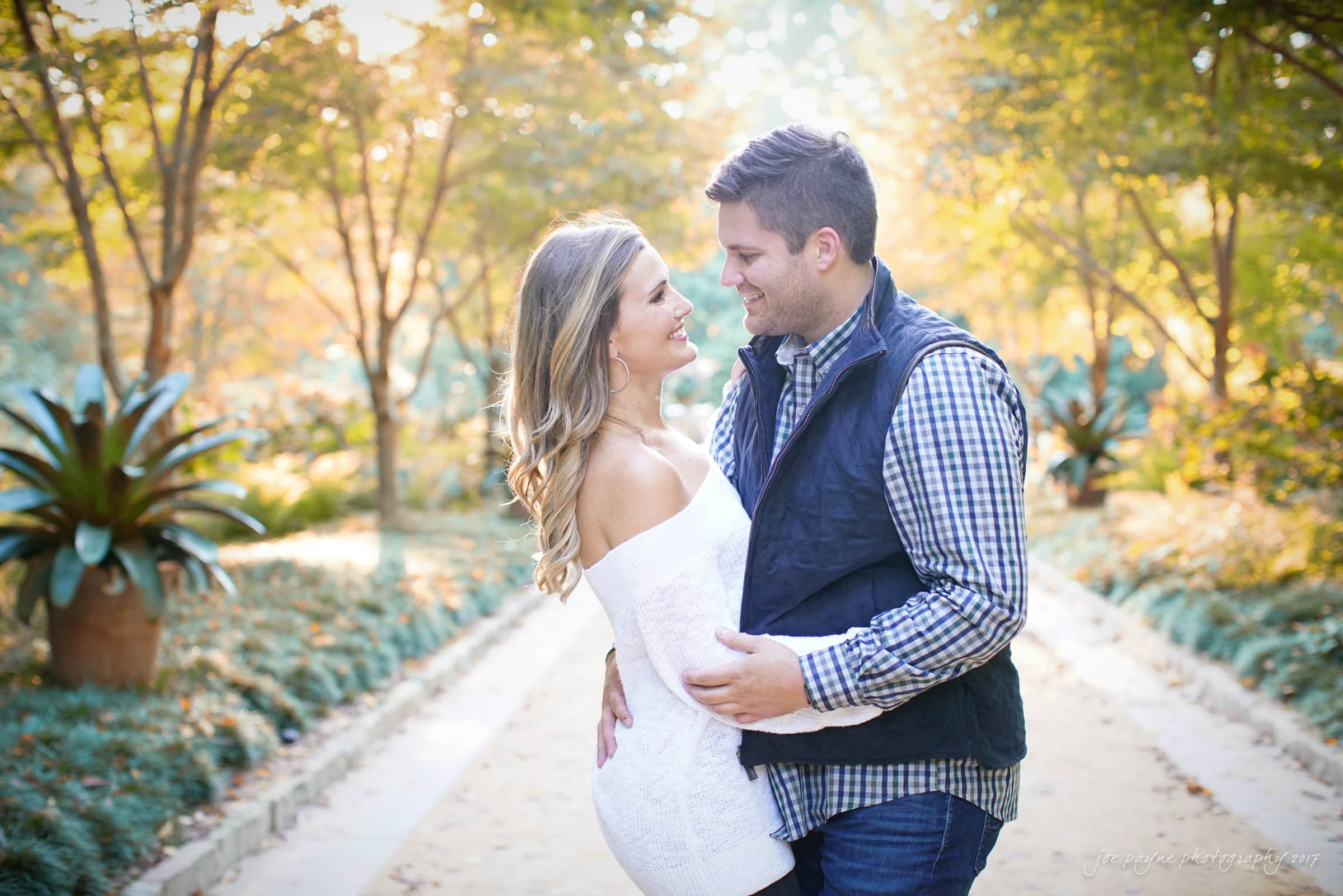 A couple stands closely, smiling at each other in a sunlit garden. The woman is wearing a white dress, and the man is in a plaid shirt with a dark vest. Lush greenery and soft sunlight create a romantic atmosphere in the background.
