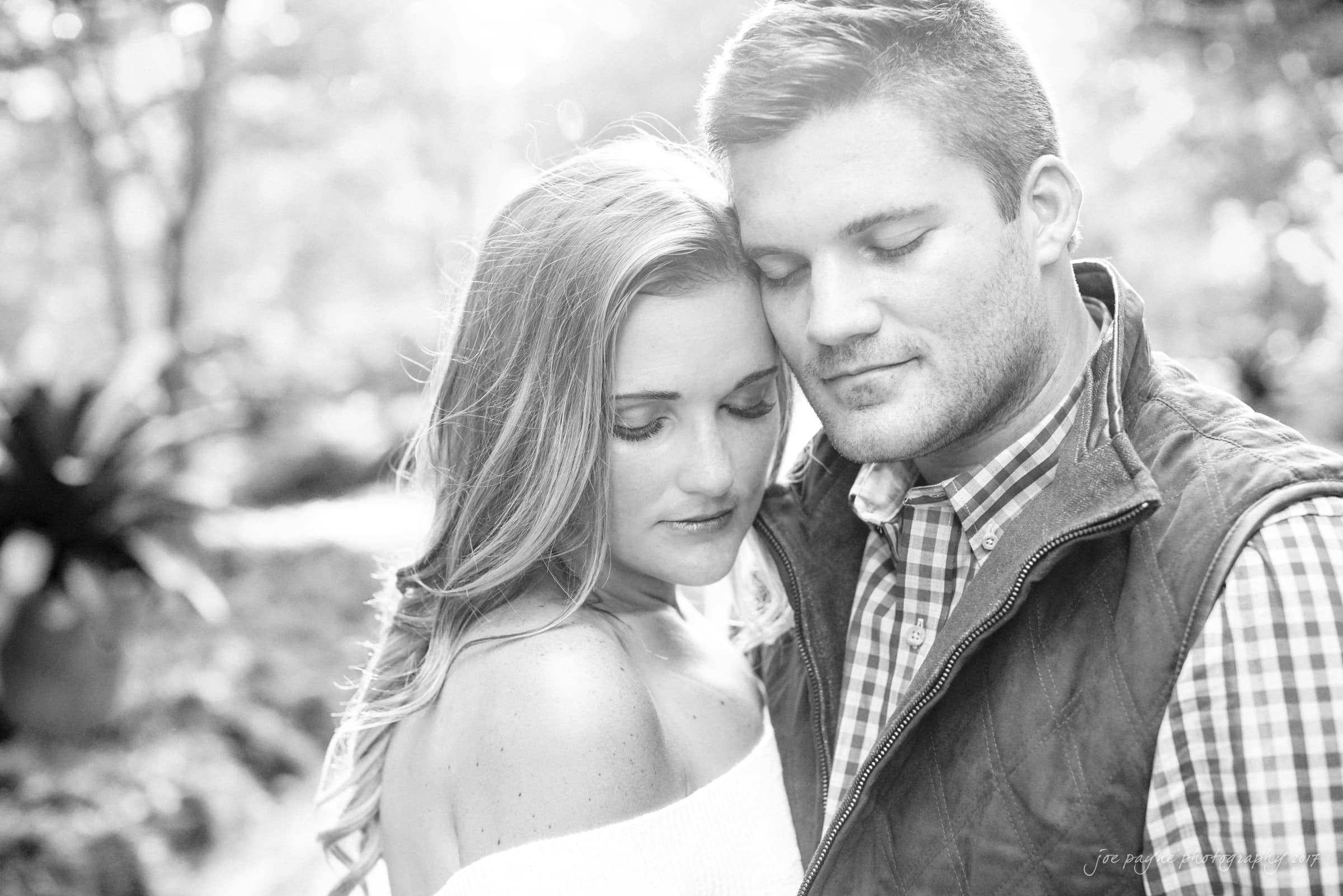 A black and white photo of a couple embracing outdoors. The woman, with long hair, looks downward while the man, wearing a checkered shirt and vest, closes his eyes and leans his head against hers. Sunlight filters through the trees, creating a soft glow.