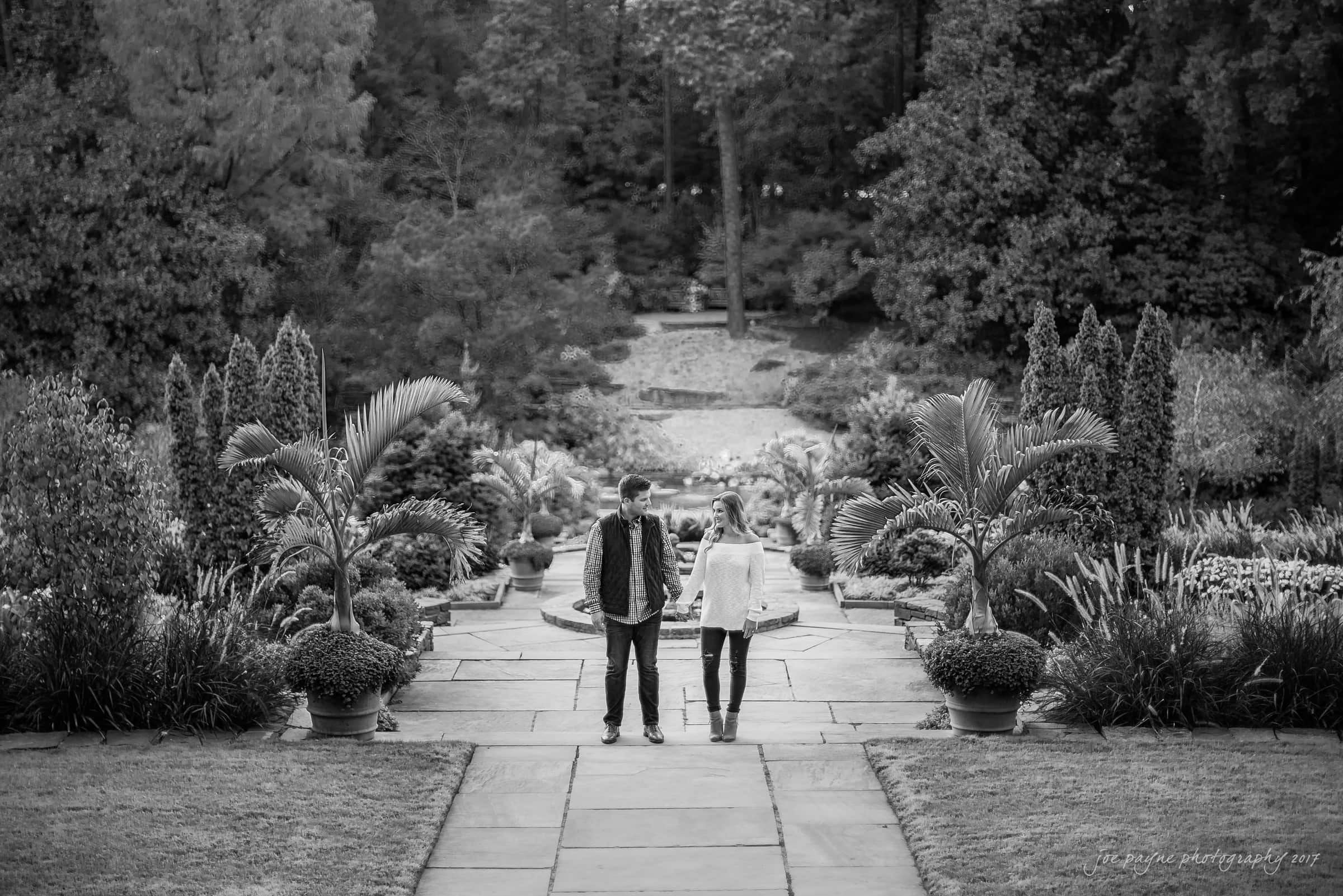 A black and white image of a couple walking hand in hand along a stone pathway in a lush garden. Tall trees and various plants surround them, creating a serene and peaceful atmosphere. The scene captures a moment of togetherness and tranquility.
