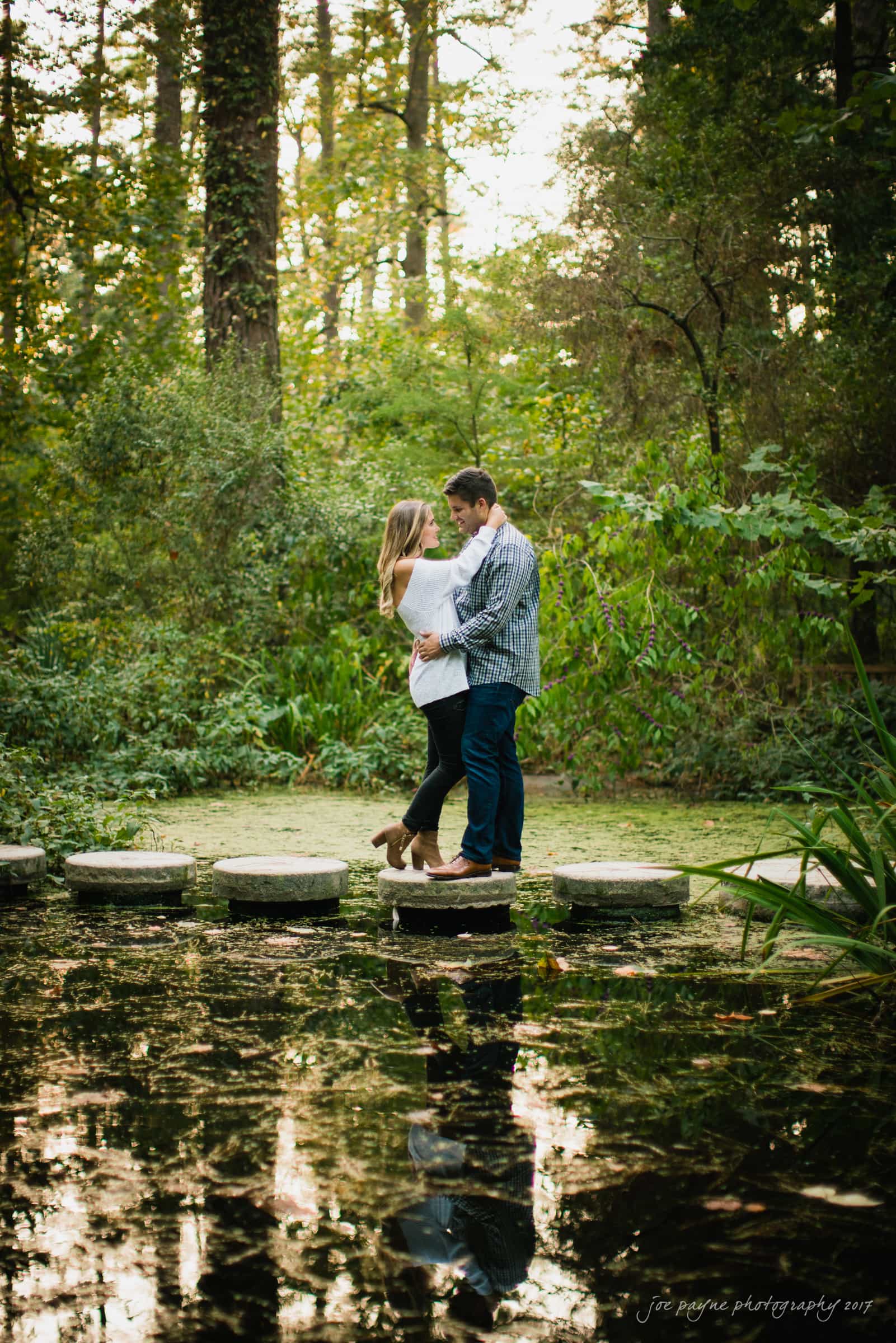 A couple embraces romantically while standing on stepping stones over a tranquil pond. The surrounding greenery and tall trees provide a serene, natural backdrop, with the water reflecting their image.