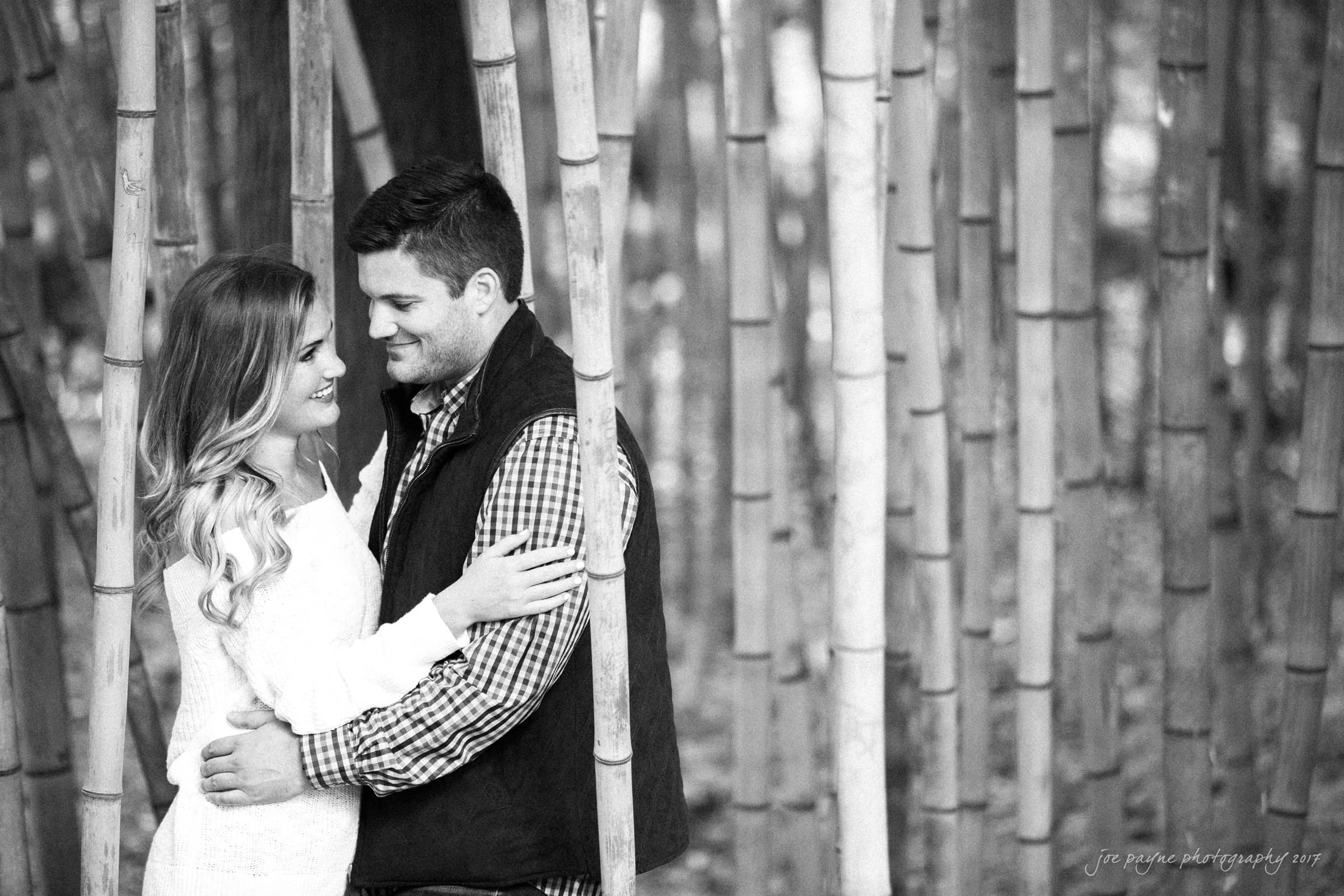A couple stands closely in a bamboo forest, smiling and gazing into each others eyes. The woman wears a light-colored jacket, and the man wears a checkered shirt with a dark vest. The background features tall bamboo stalks. The image is in black and white.