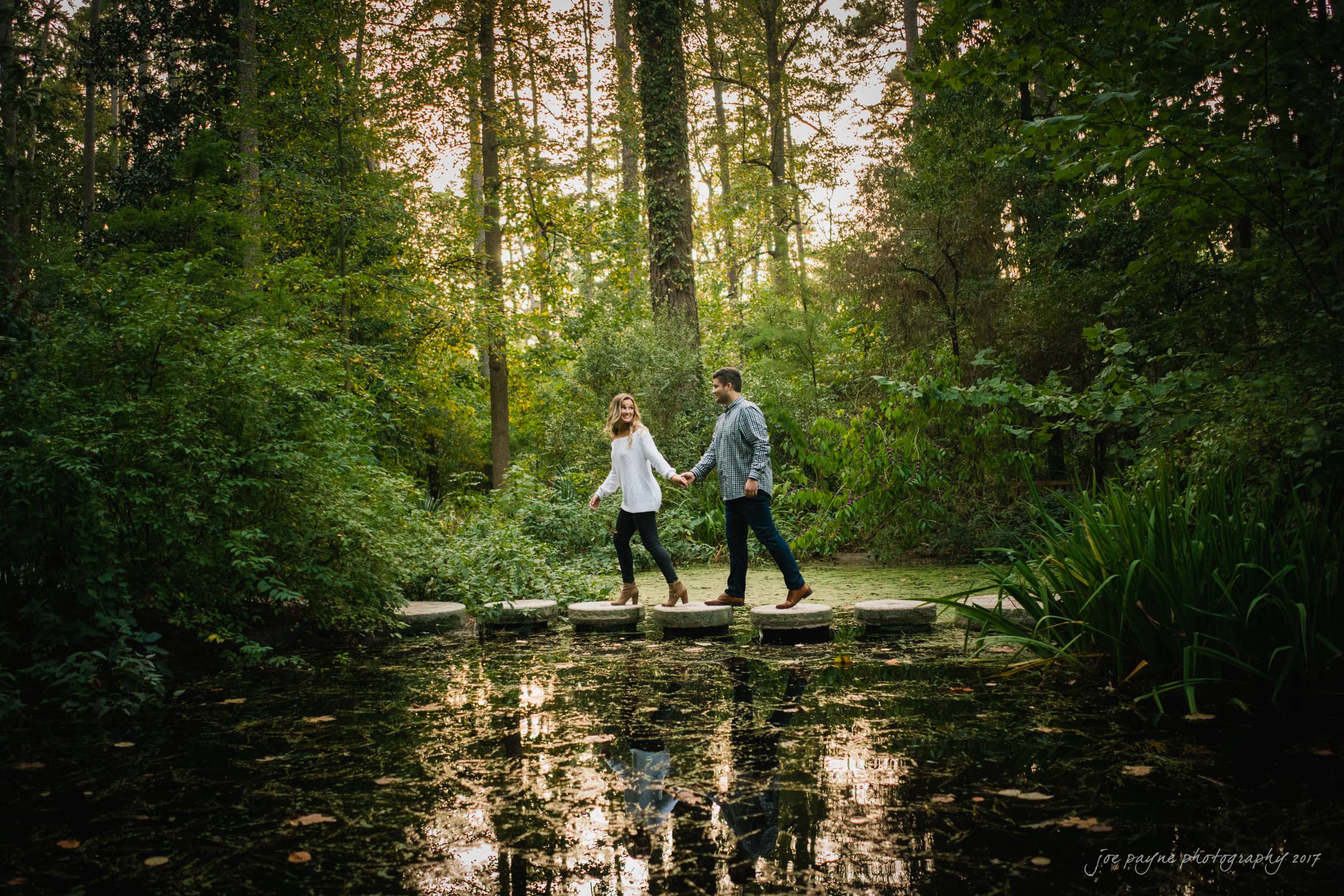 A couple holding hands, walking on stepping stones across a pond surrounded by lush greenery. The setting sun casts a warm glow through the trees, reflecting on the water.