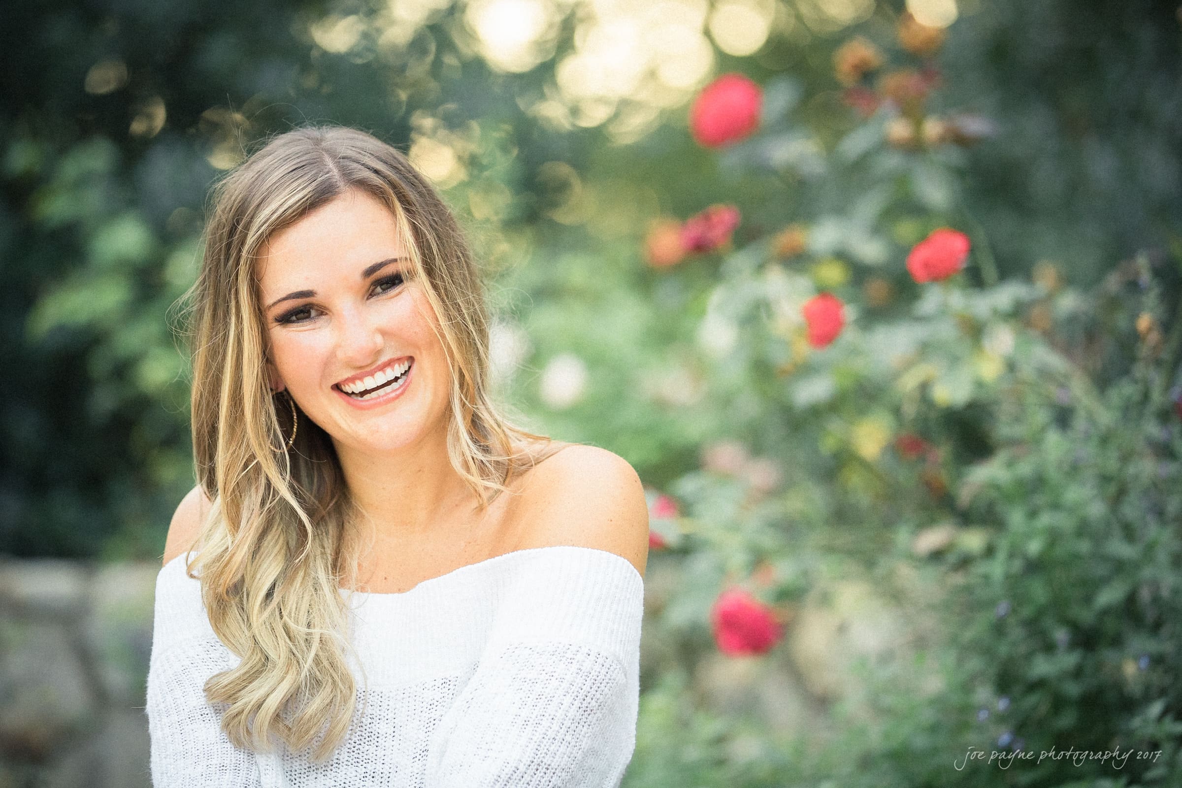 A woman with long blonde hair smiles while wearing a white off-shoulder sweater. She is sitting outdoors with a background of blurred greenery and pink flowers. The sunlight creates a warm and inviting atmosphere.