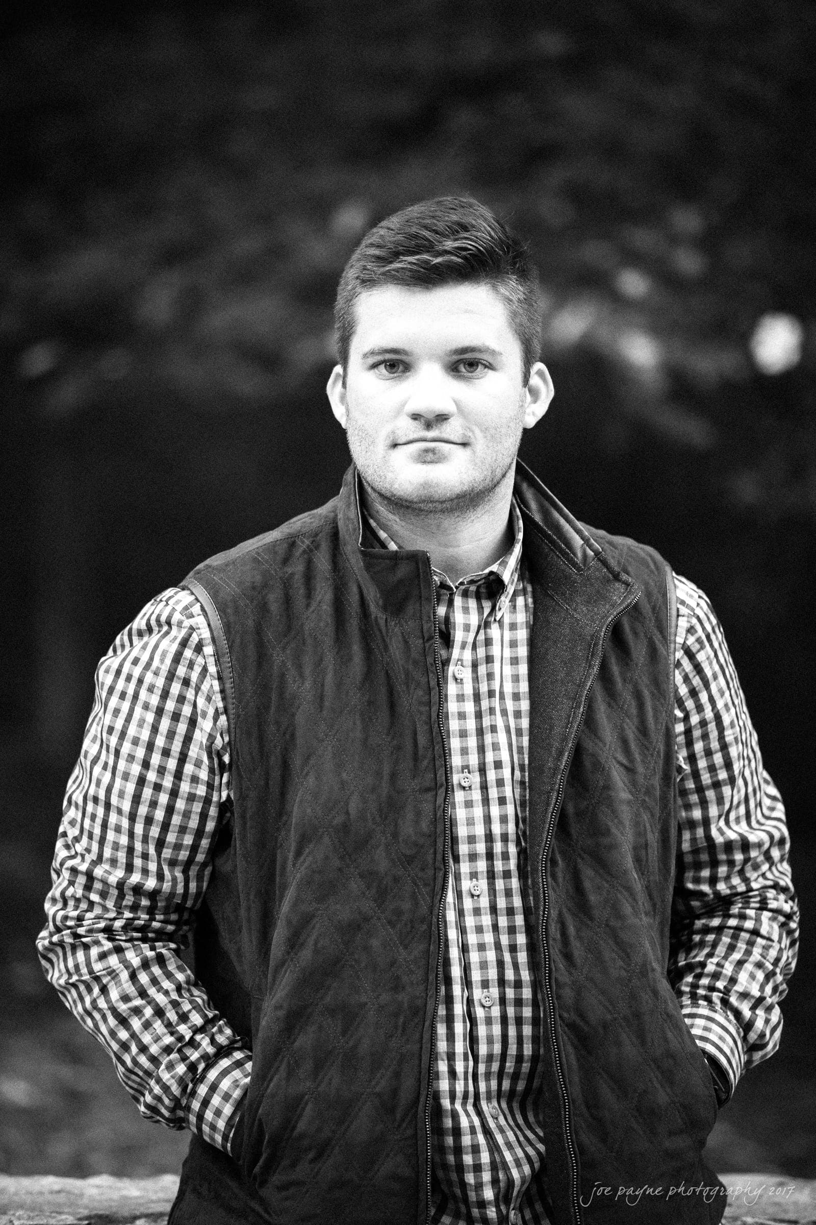 A man stands outdoors, leaning slightly against a railing. He is wearing a checkered shirt and a quilted vest. The setting is dimly lit, with a dark, out-of-focus background, suggesting trees or foliage. He has his hands in his vest pockets.