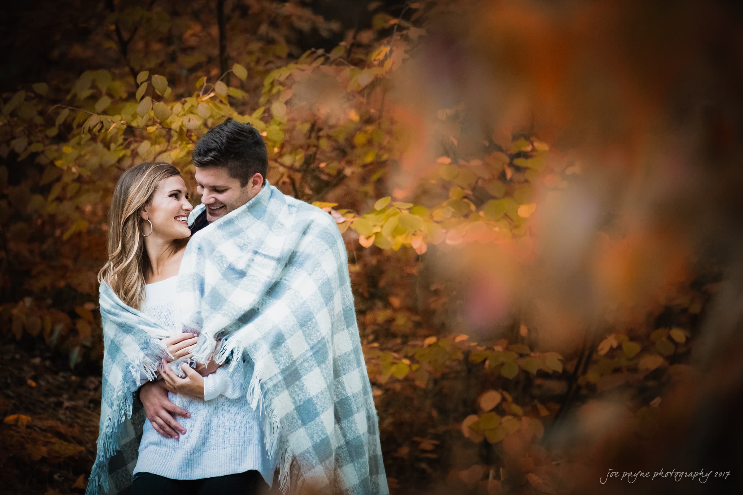 A couple stands wrapped in a checkered blanket, smiling at each other amidst autumn foliage. The woman wears a white sweater, and the man is in a dark sweater, creating a cozy and warm atmosphere.