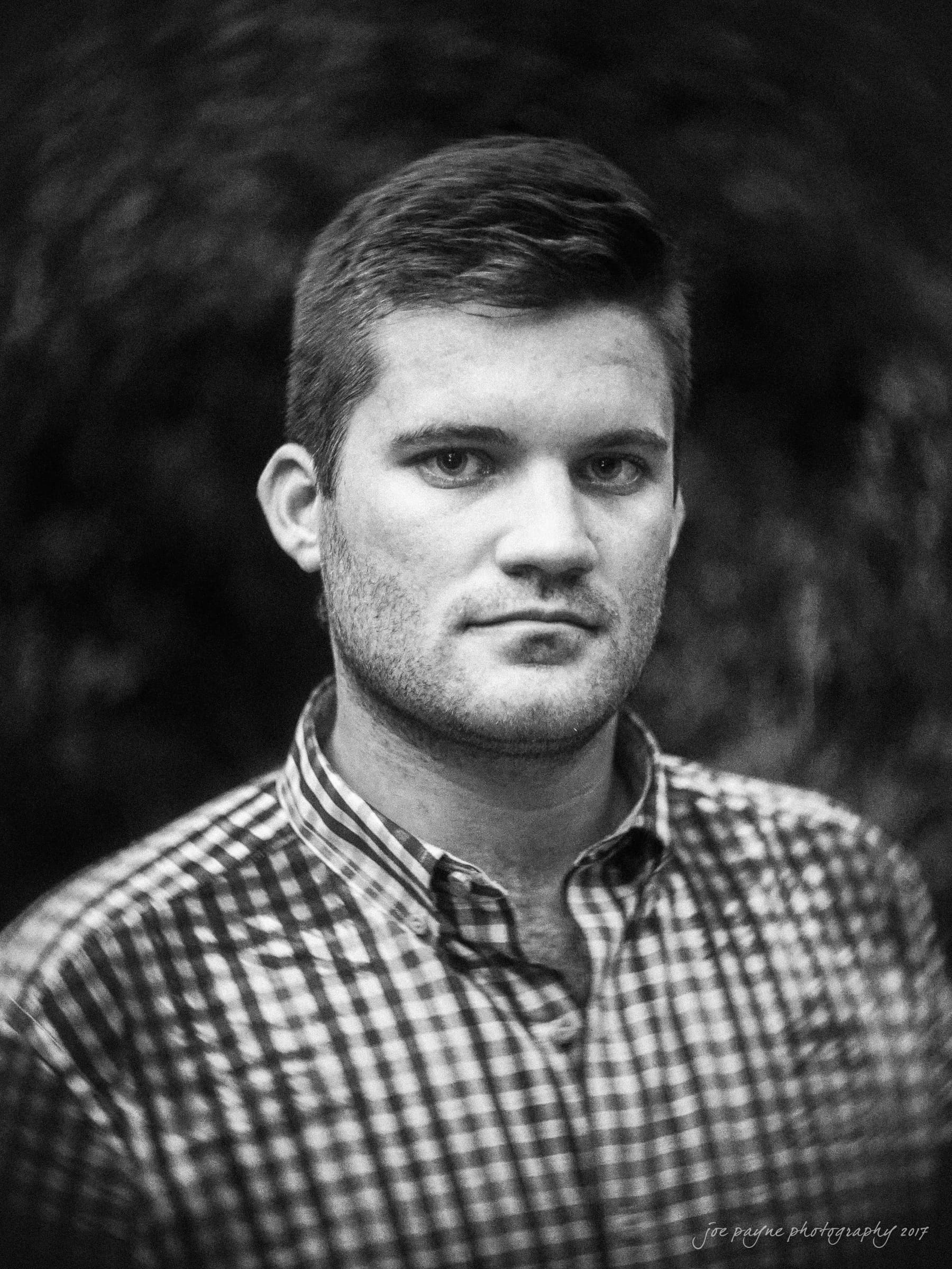 A black and white portrait of a person with short hair, wearing a plaid shirt. The background is blurred, focusing on the persons expression.