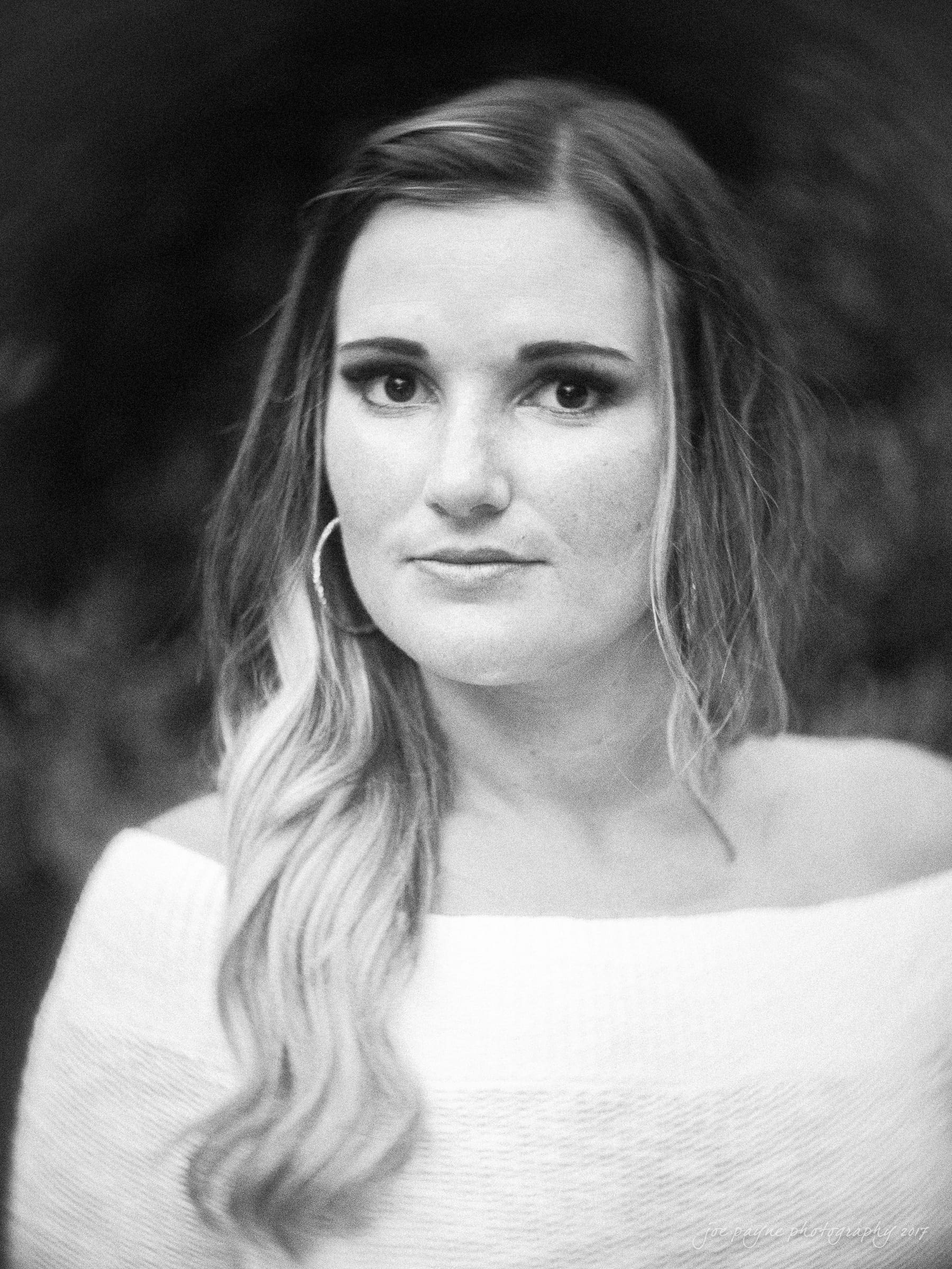 Black and white portrait of a woman with long hair wearing a strapless top and hoop earrings. She gazes directly at the camera with a neutral expression, set against a blurred background.