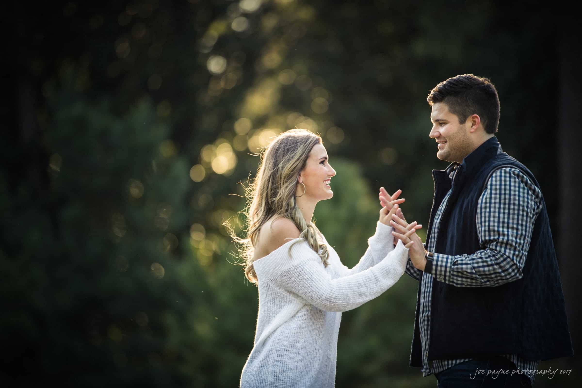 A couple stands facing each other in front of a blurred natural background. The woman wears a white sweater, and the man wears a black vest over a plaid shirt. They appear to be in a playful and affectionate moment, holding hands.
