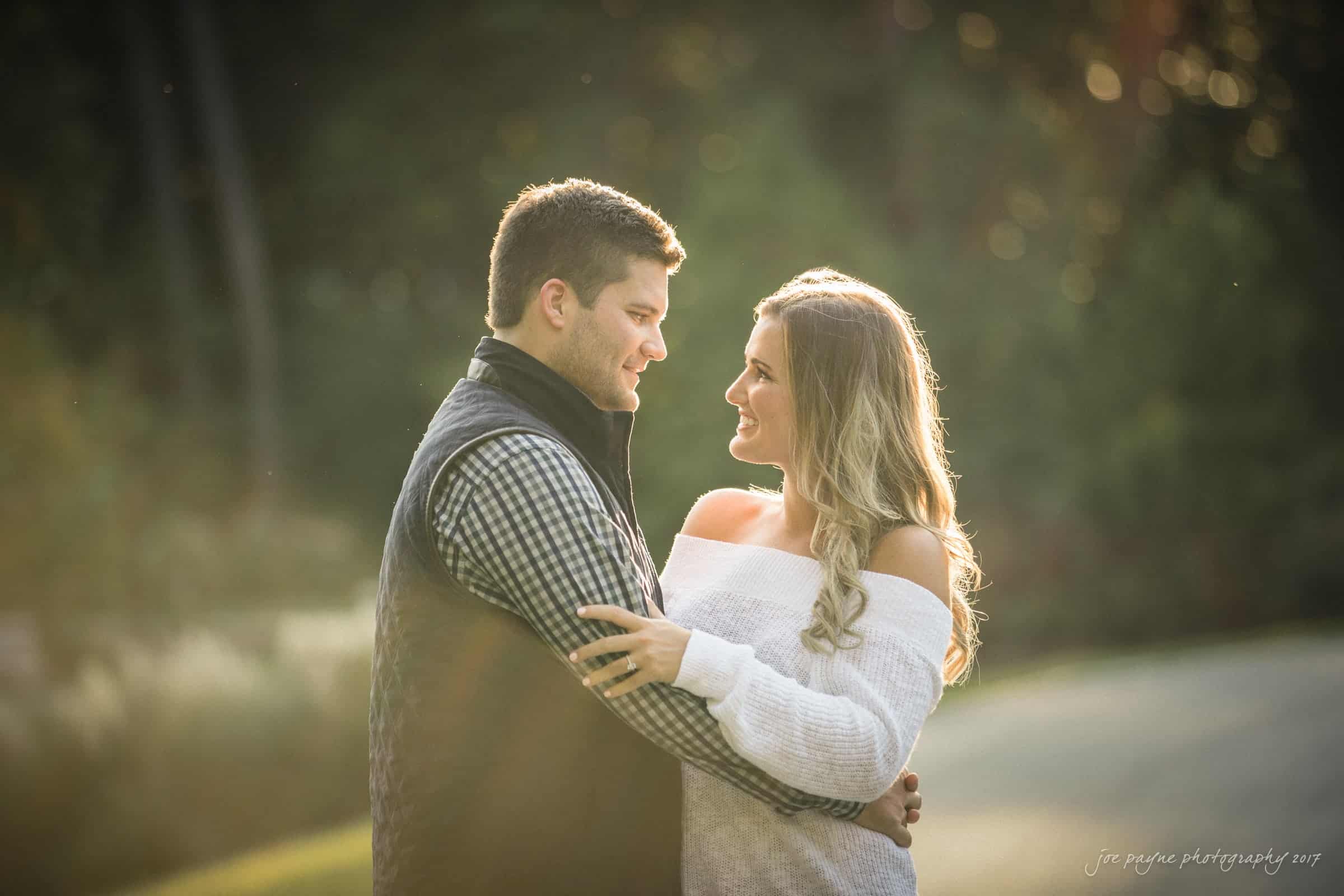 A couple stands outdoors, embracing and smiling at each other. The woman wears a white off-shoulder sweater, and the man wears a checkered shirt with a vest. Sunlight filters through the trees in the background, creating a warm, serene atmosphere.