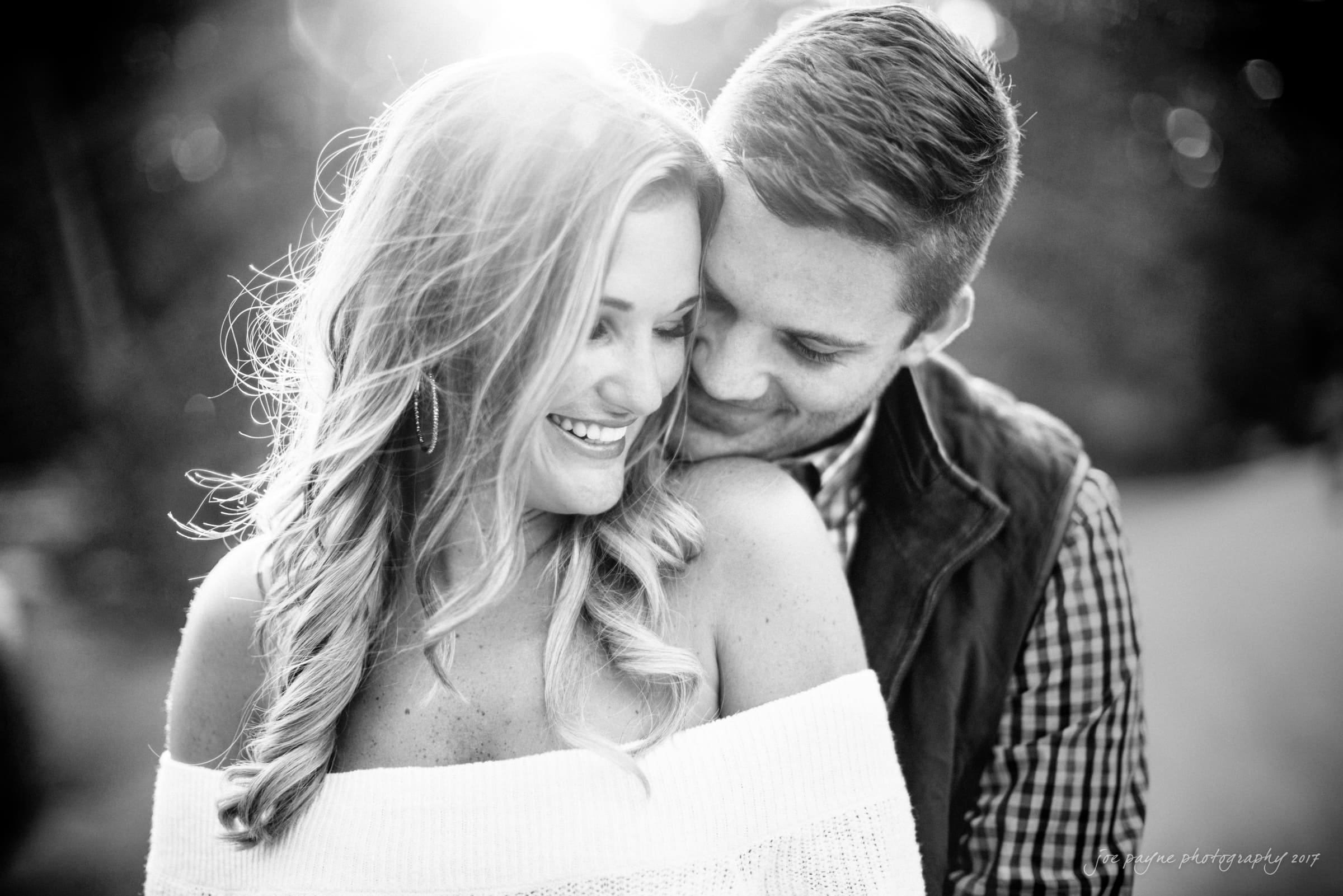 A black-and-white photo of a smiling couple at Duke Gardens. The woman, in an off-shoulder sweater, looks down as the man stands behind her, gently nuzzling her shoulder—sunlight softly illuminating this tender engagement photography moment.
