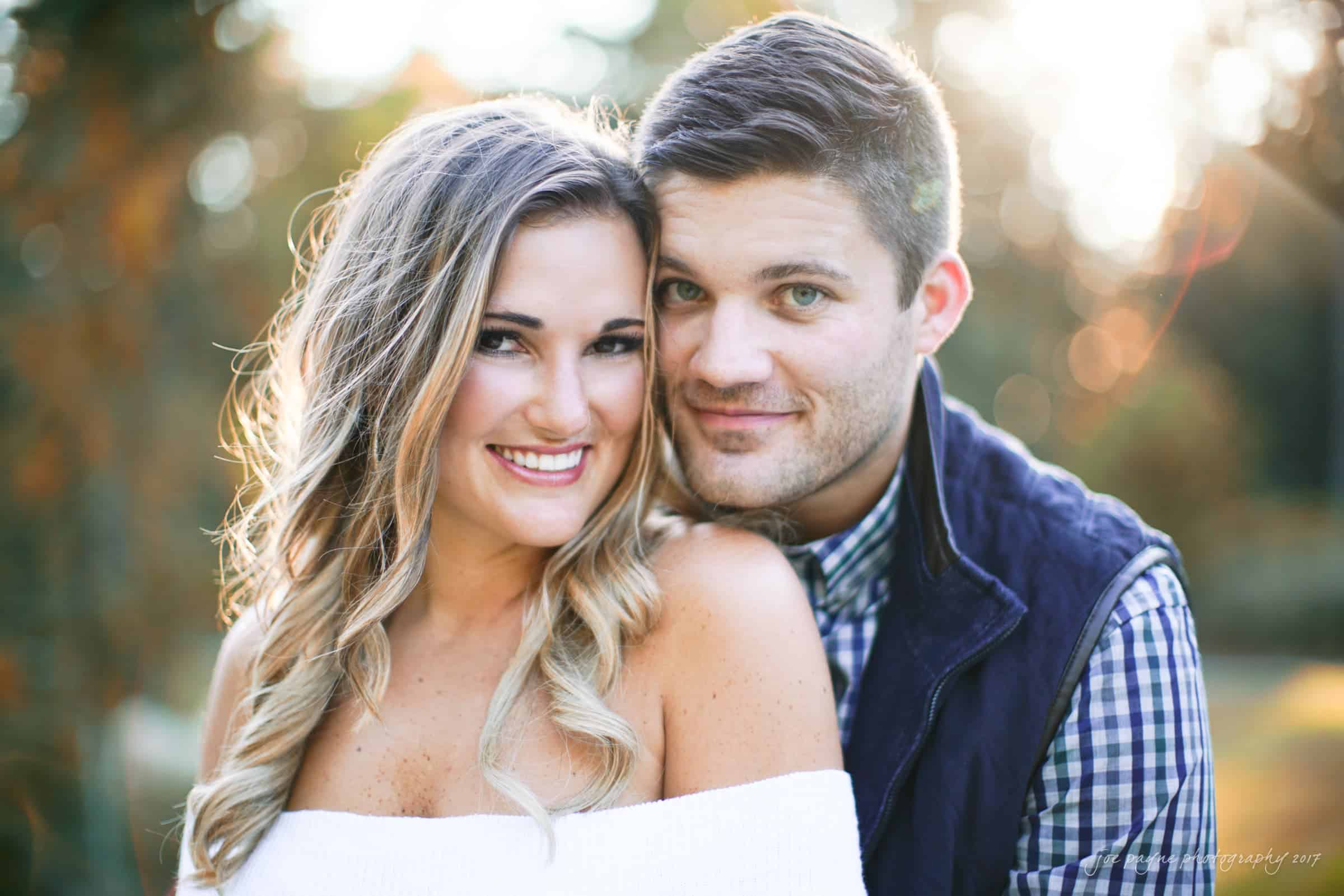 A smiling couple poses outdoors in soft, natural light. The woman wears an off-shoulder top, and the man has a checkered shirt and dark vest. Blurred trees and sunlight create a warm, inviting background.