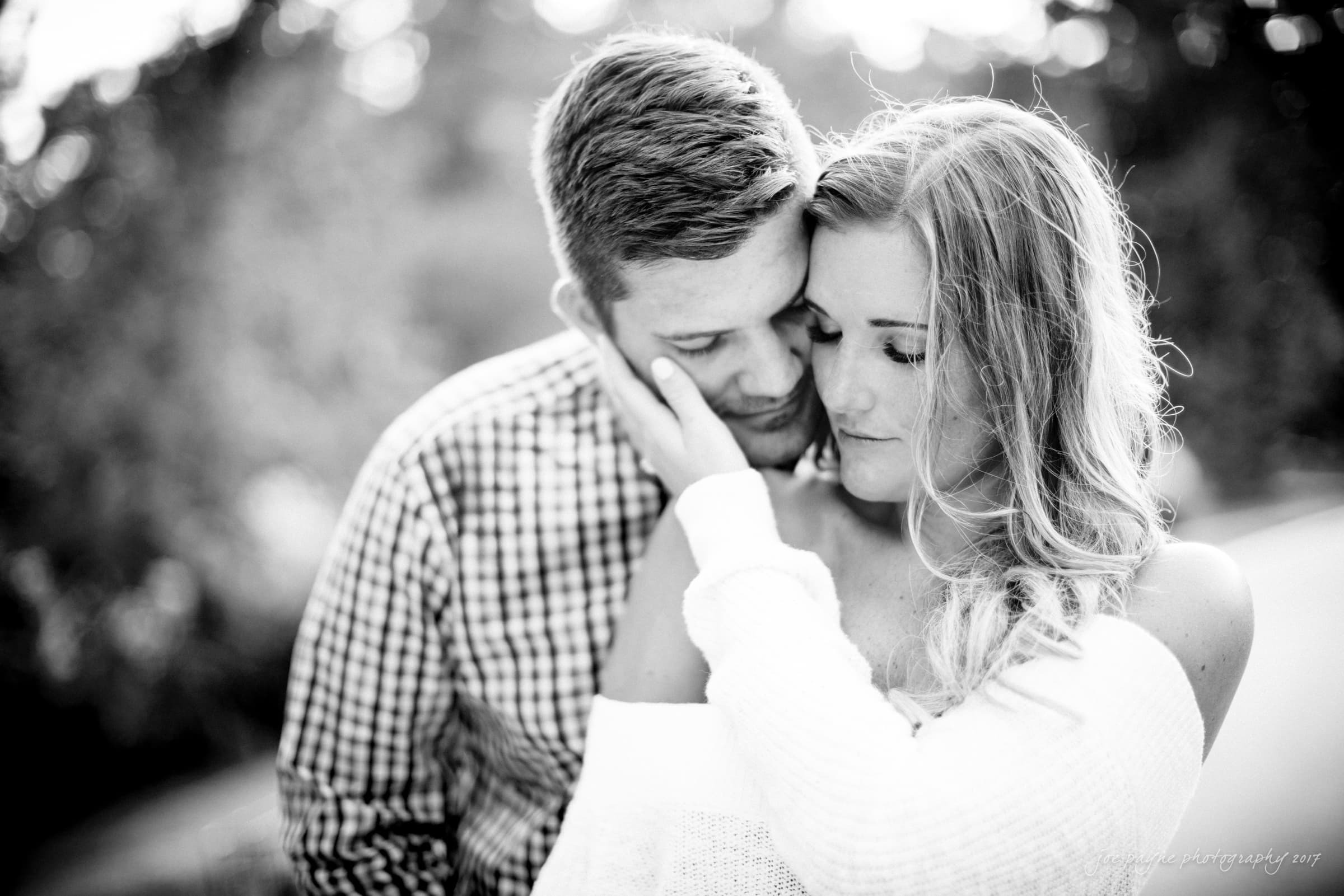 A black and white photo of a couple embracing. The man gently rests his head on the womans forehead, while she wraps a hand around his neck. They both appear content and serene, surrounded by blurred, natural scenery.