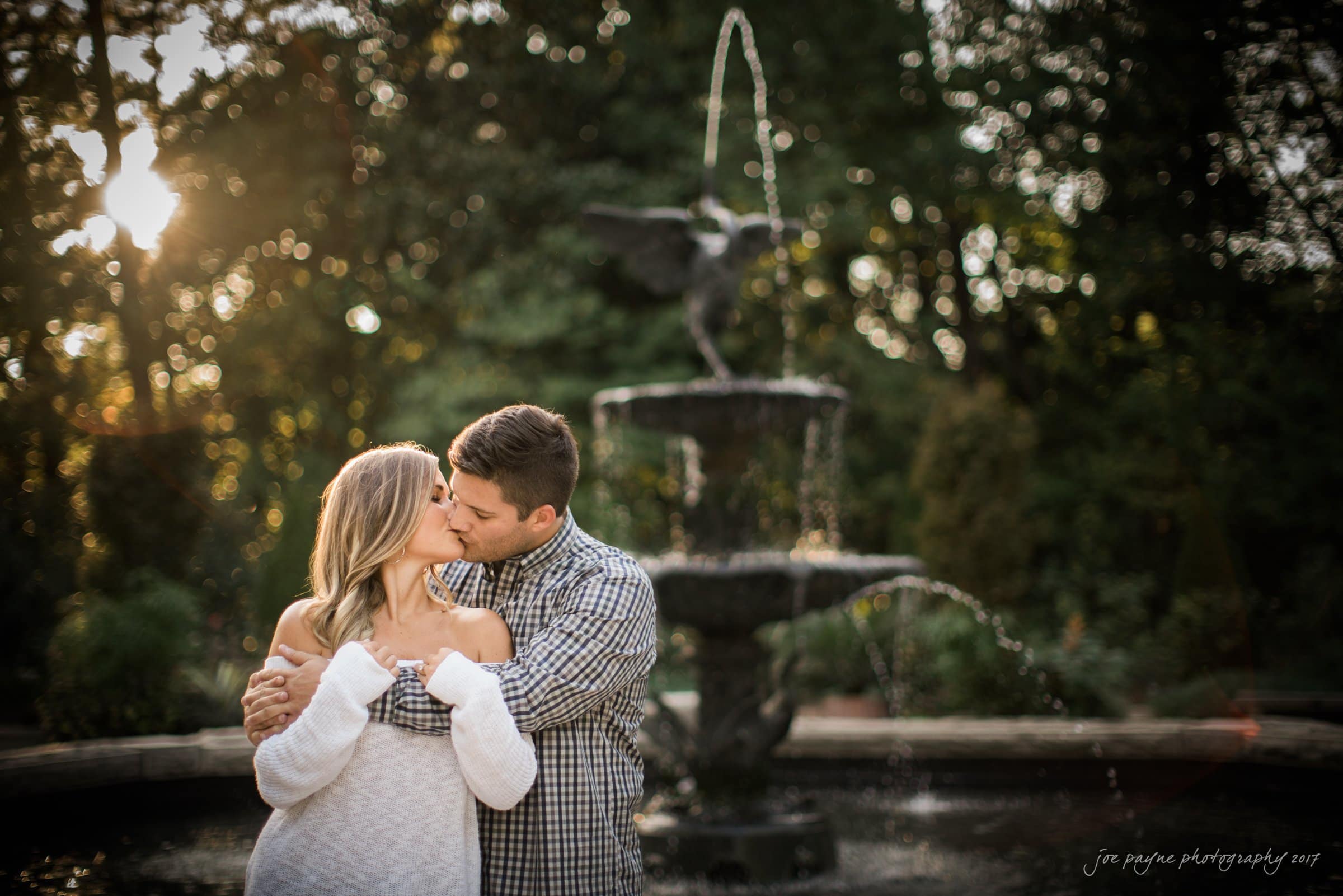 A couple embraces and kisses in front of a picturesque fountain, surrounded by lush greenery. The sun sets in the background, casting a warm glow and creating a romantic atmosphere.